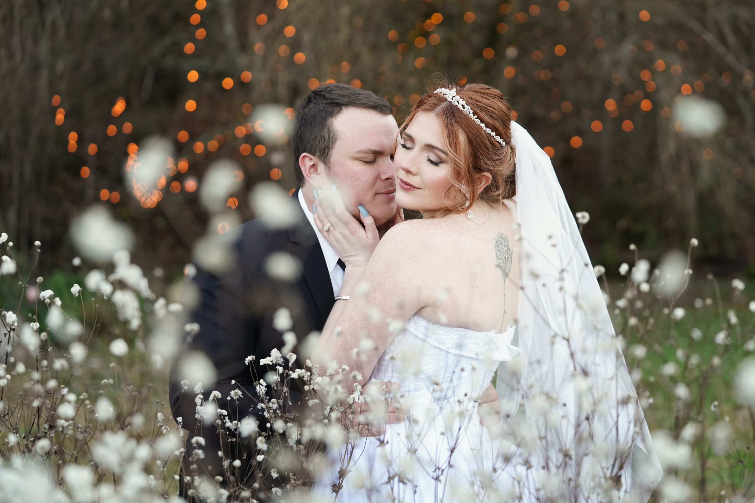 Bride lovingly embracing her groom in front of fluffy white fall seeds near a willow tree with warm fairy lights