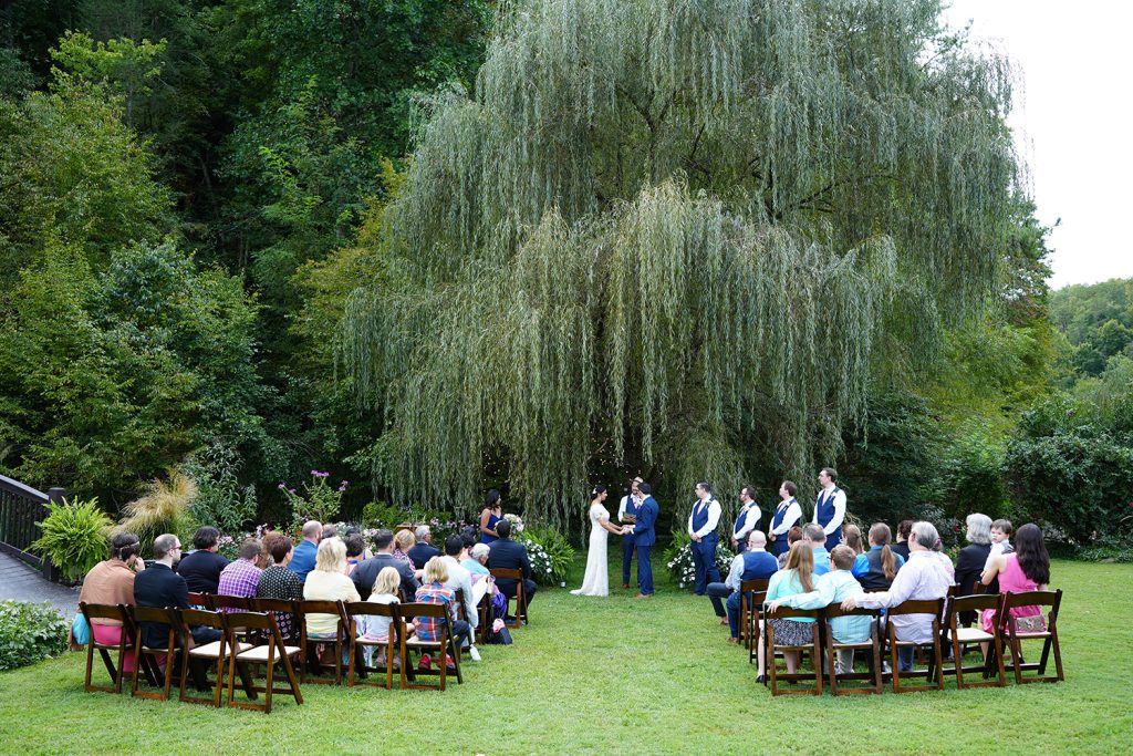 Wedding ceremony in Tennessee under a willow tree with several guests sitting in dark wooden folding chairs