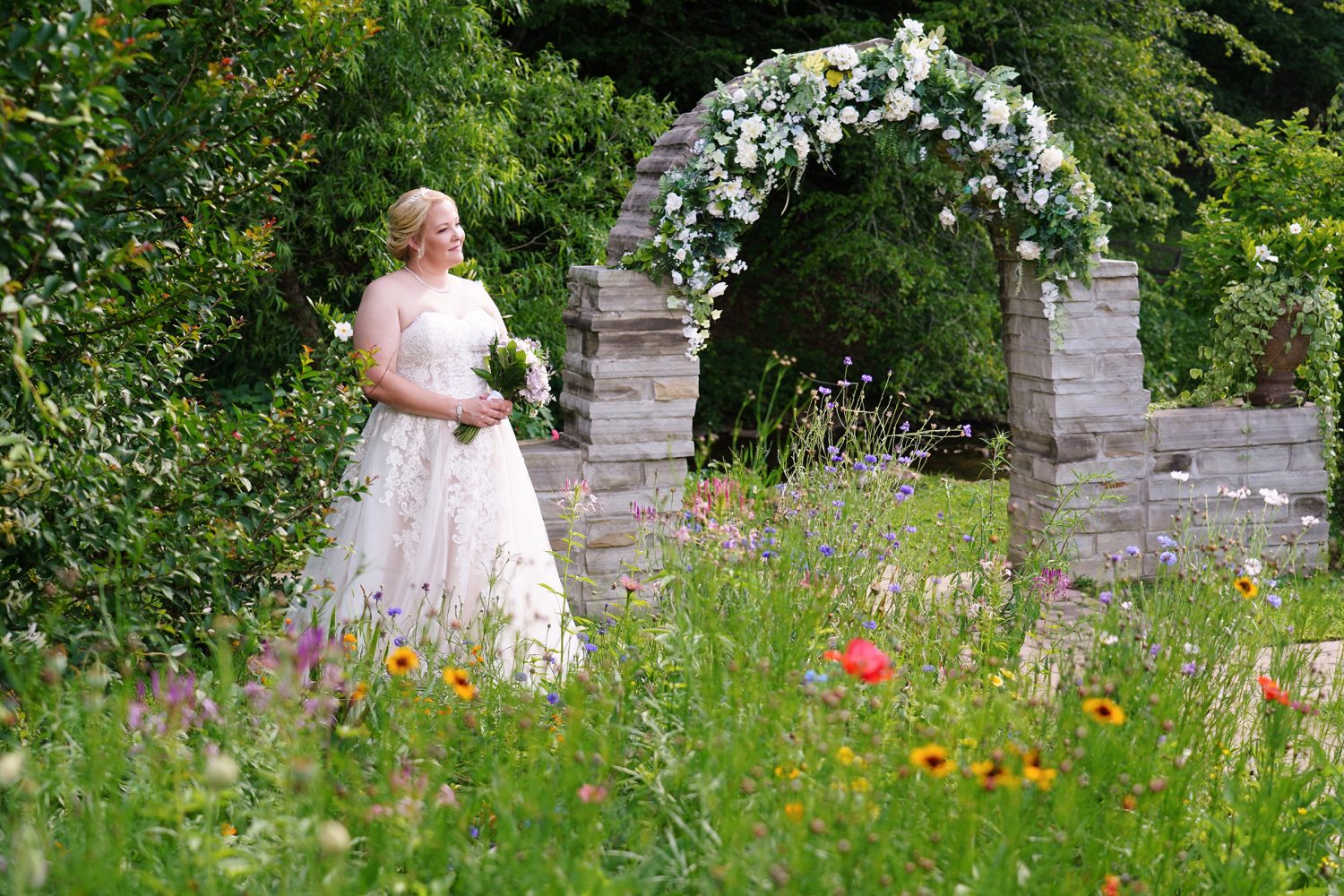 Bride by a stone wedding arch decorated with white flowers in a spring wildflower garden