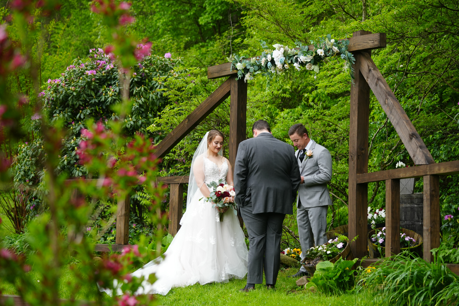 Wedding ceremony at a wooden arbor in the spring with foliage all around and red flowers in bloom in the foreground