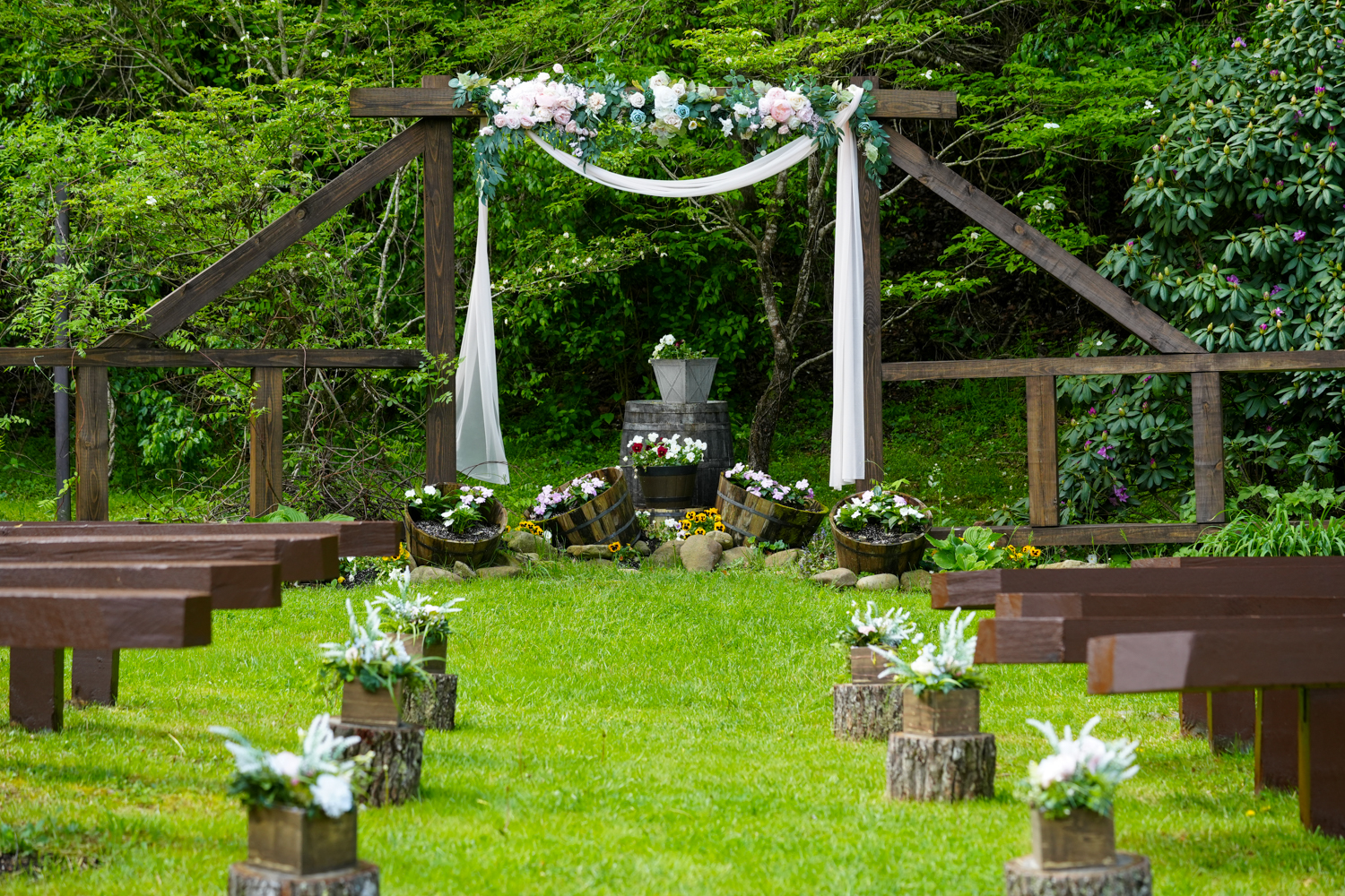 Wooden wedding arbor called Creekside Ridge at the Honeysuckle Hills wedding venue with dark brown benches and flowers in wooden vases sitting on top of stumps leading to a pink drape and lings moments garlands on the arbor