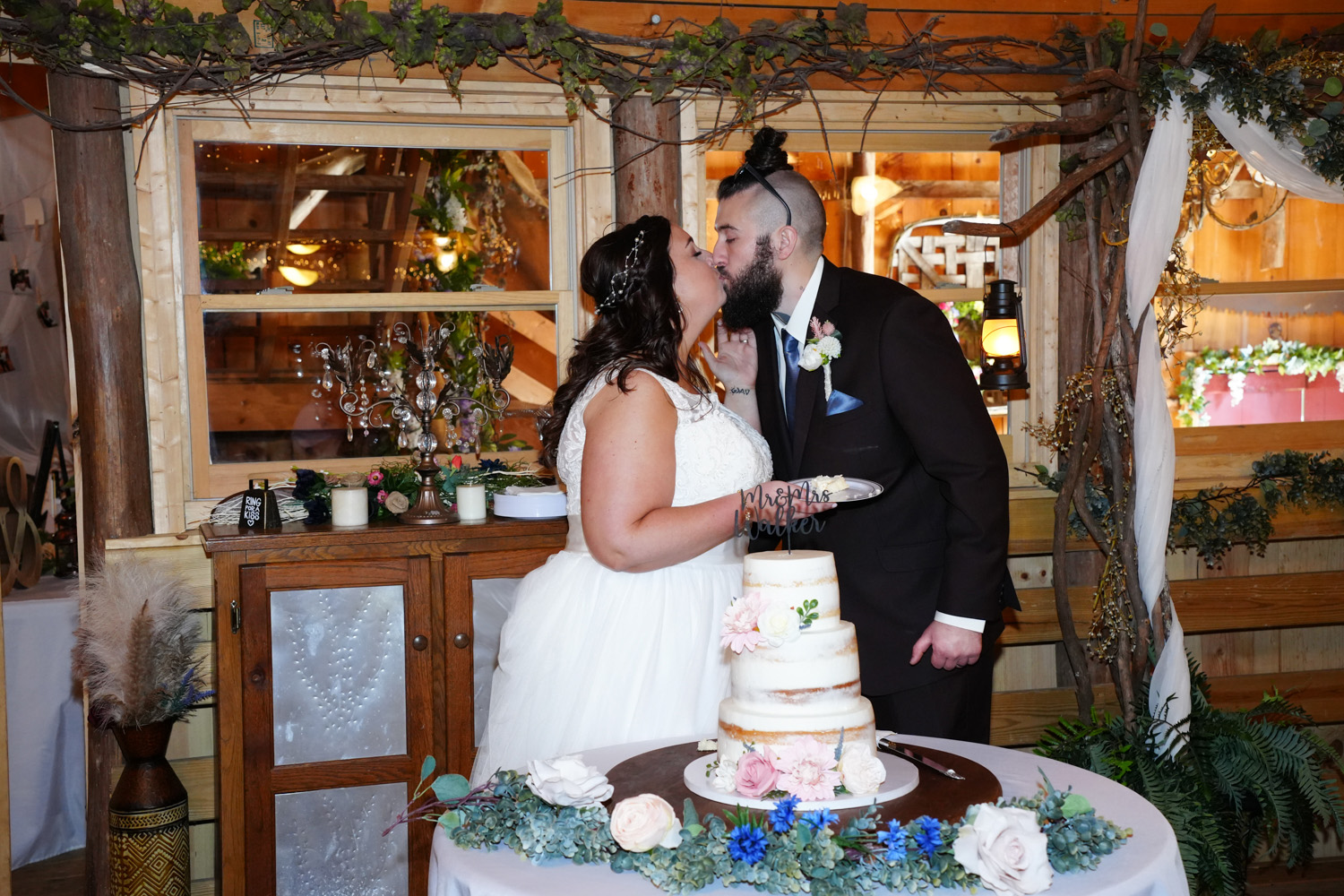 Bride and groom kissing in front of their wedding cake in a barn wedding venue with country chic decor