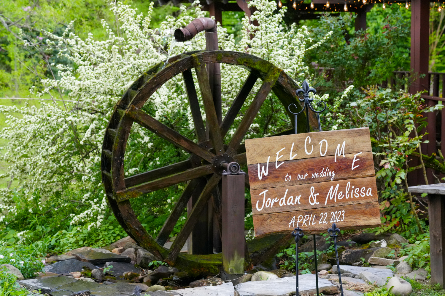 white pyracantha in bloom by a water wheel at the Honeysuckle Hills wedding venue in Pigeon Forge with a Welcome to our wedding sign on an easel