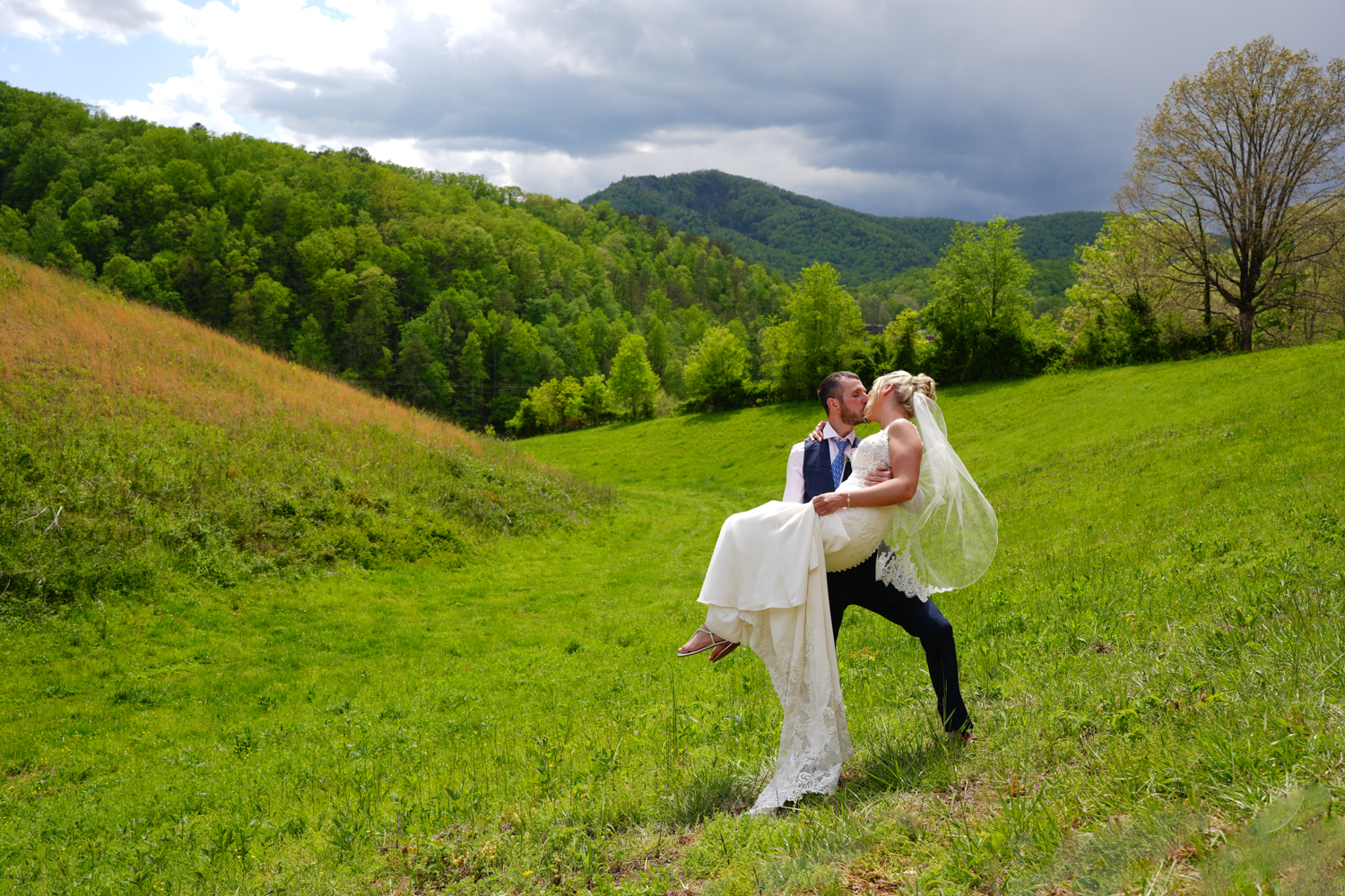 Groom holding his bride and kissing her as her veil blows in a meadow with a mountain view in Pigeon Forge at Honeysuckle Hills
