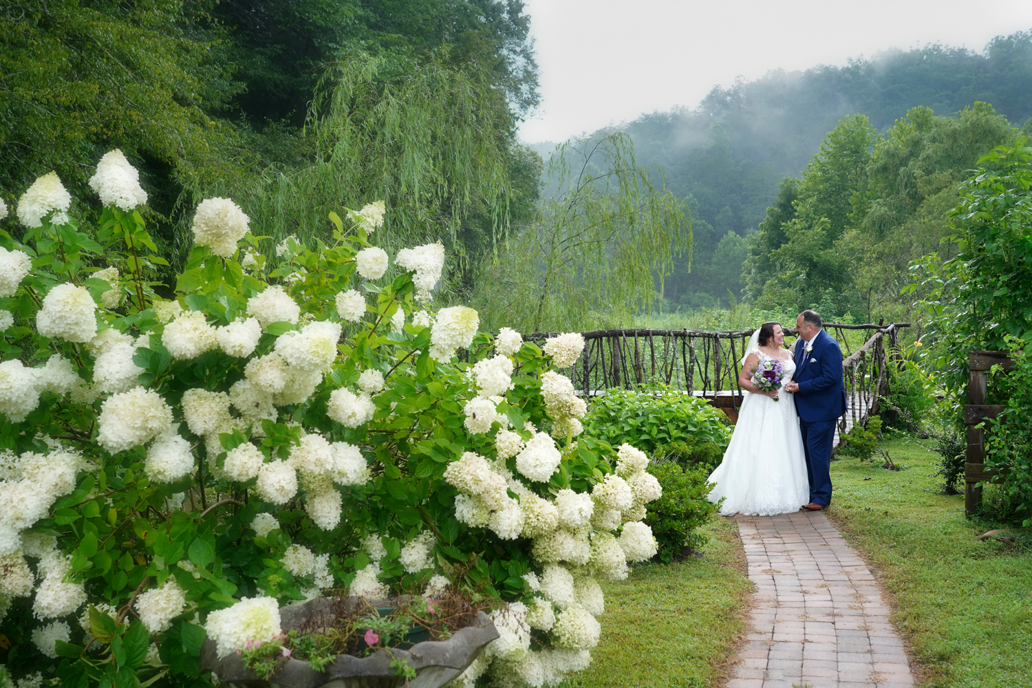 couple walking along stone path on their wedding day in a summer garden with limelight hydrangea in bloom
