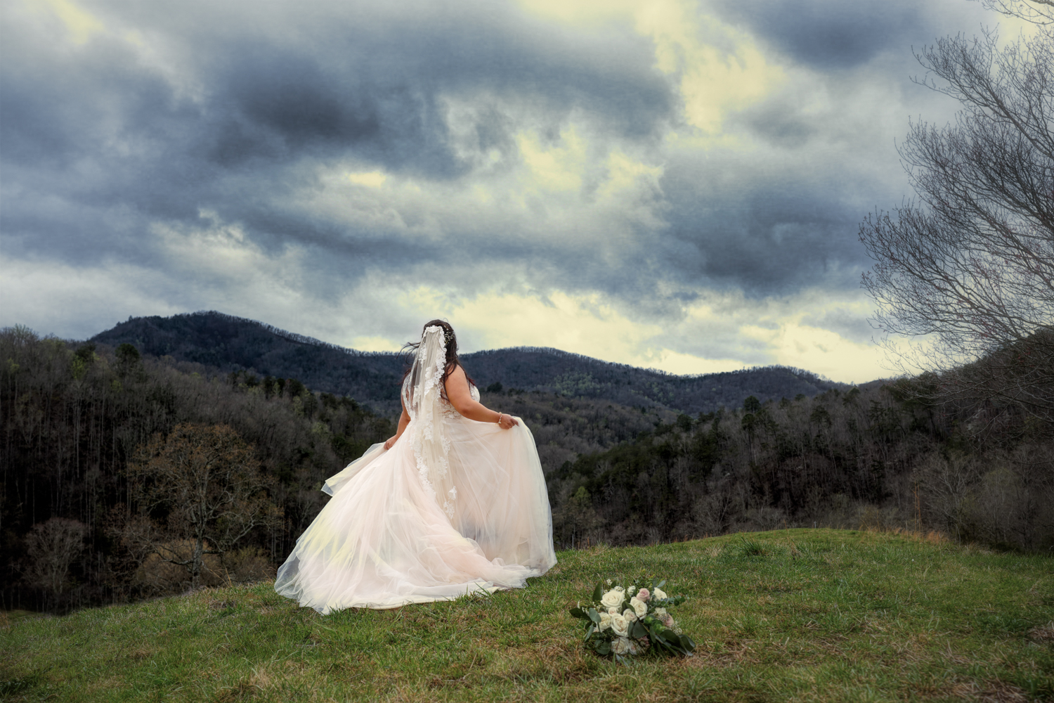 bride holding her dress as she walks in a field toward a mountain view with deep blue clouds above