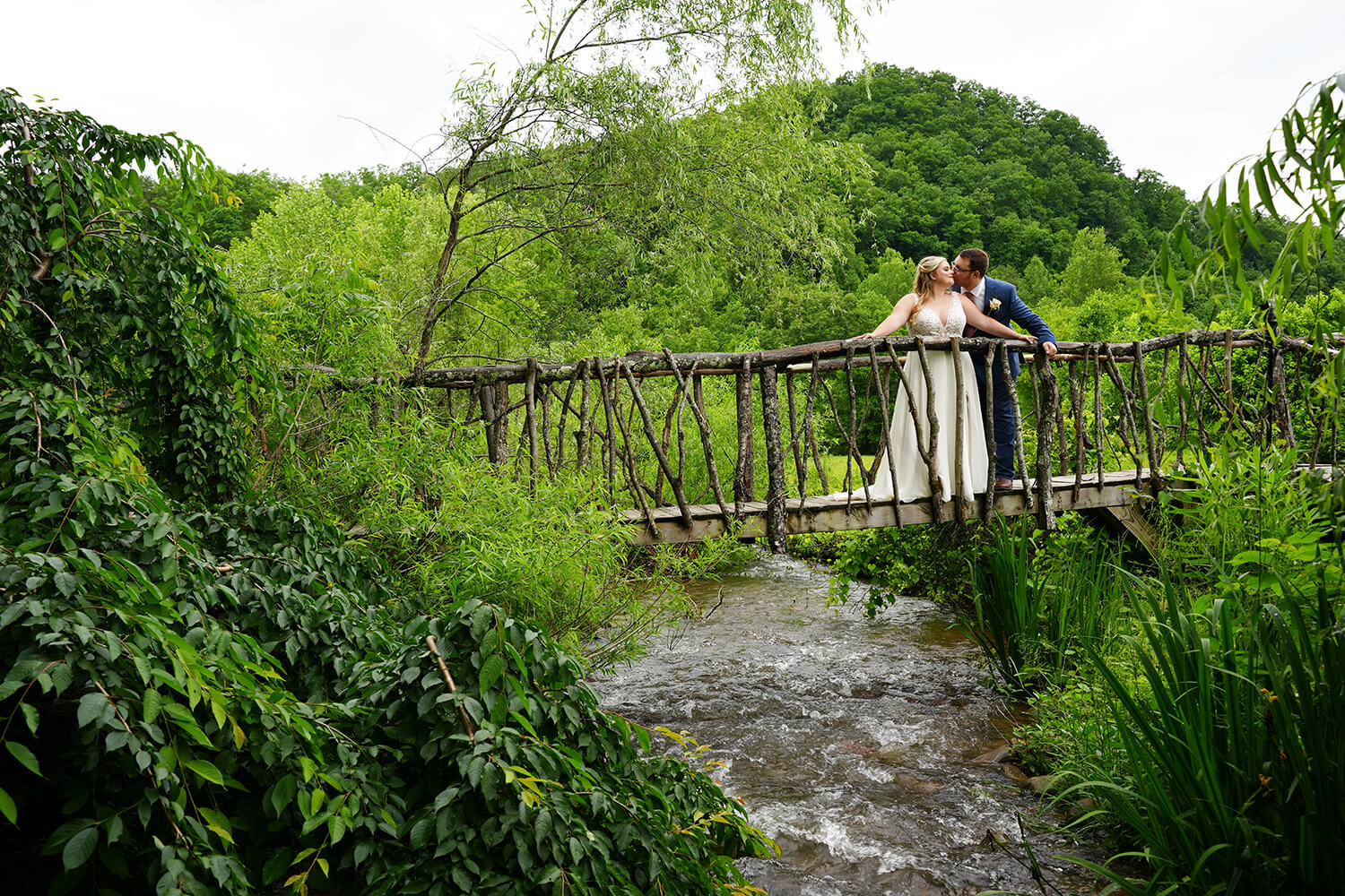 Wedding couple kissing on a tree limb hobbit bridge over a creek with a mountain ridge behind them at a wedding venue called Honeysuckle Hills in Pigeon Forge