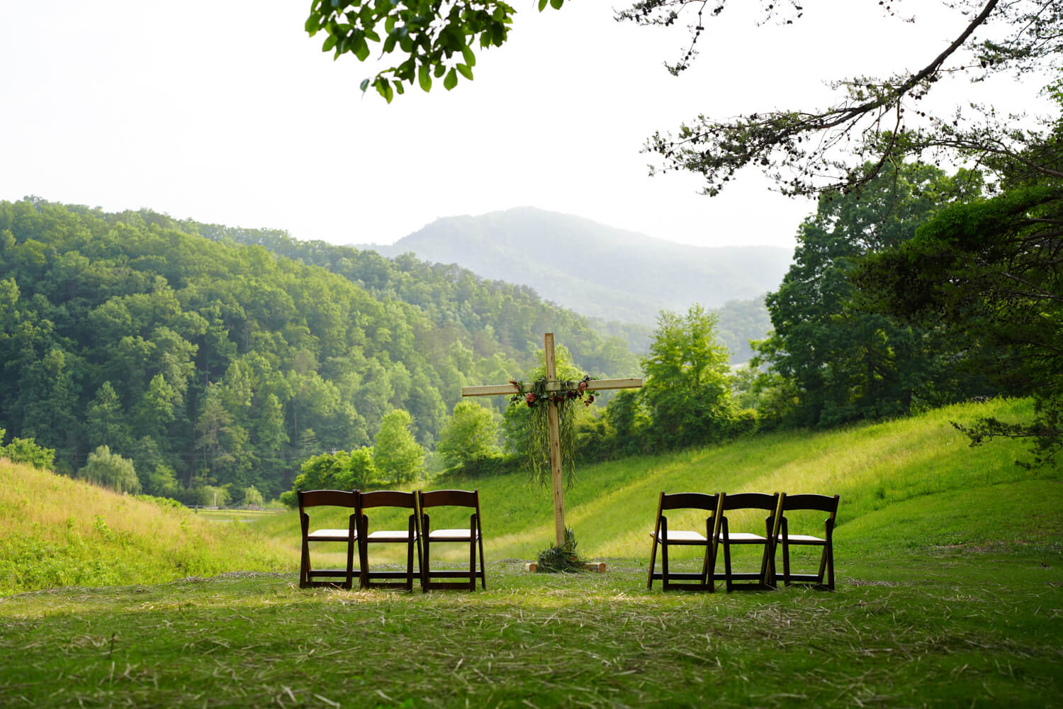 Wedding ceremony site in the Smoky Mountains with a meadow and mountain view along with dark wooden folding chairs and a tall cross decorated with vines and rust colored flowers