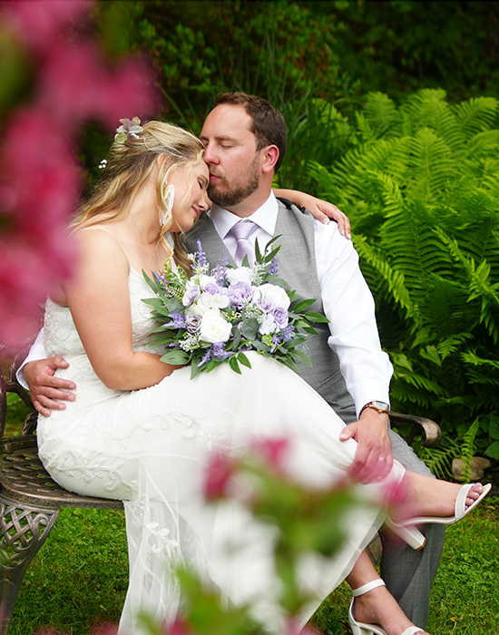 Bride sitting on her groom's lap in a garden with red flowers and green ferns as he kisses her forehead at the wedding venue where they were married