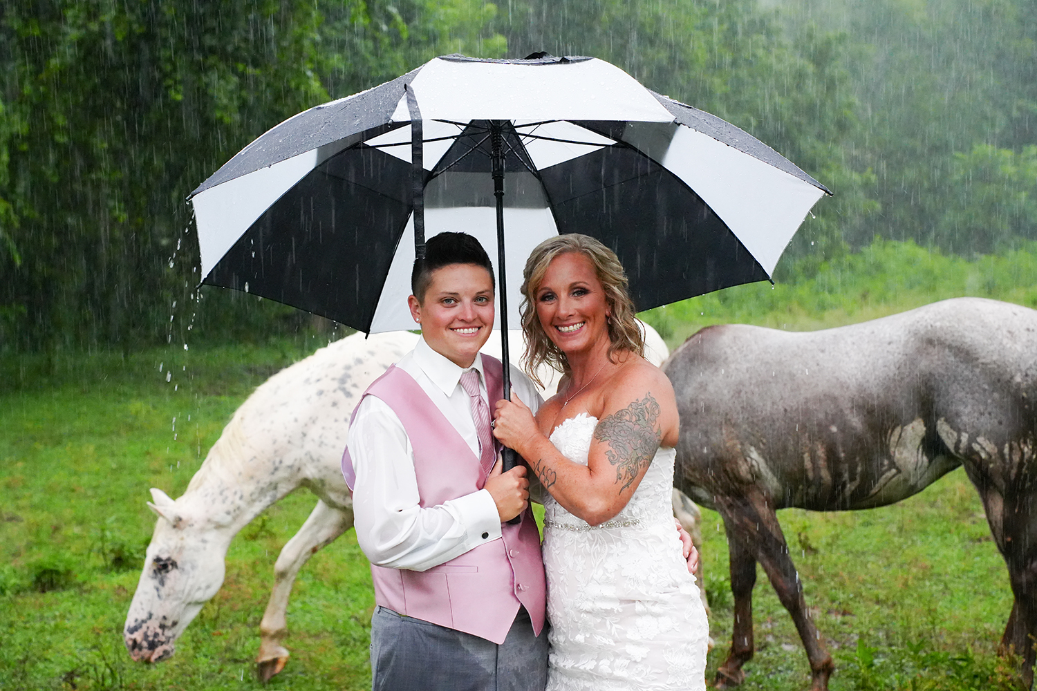 Two brides holding an umbrella in the rain in a field with two horses