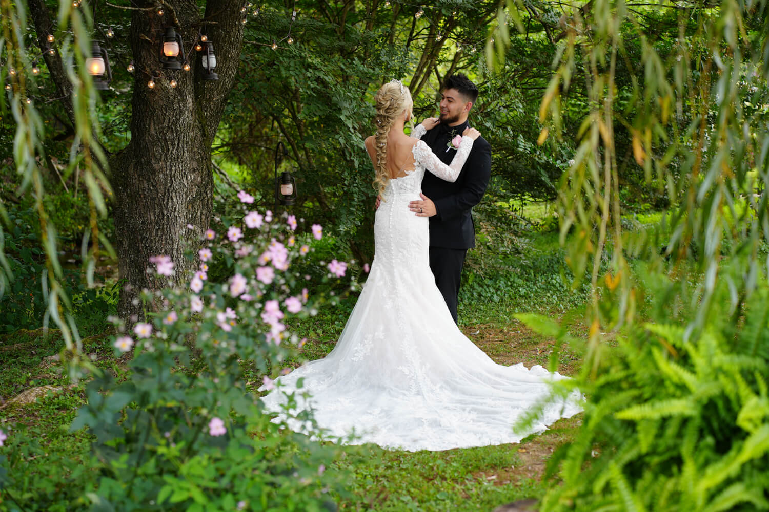 Bride with long train posing with her groom under a willow tree with lanterns and pink windflowers in bloom