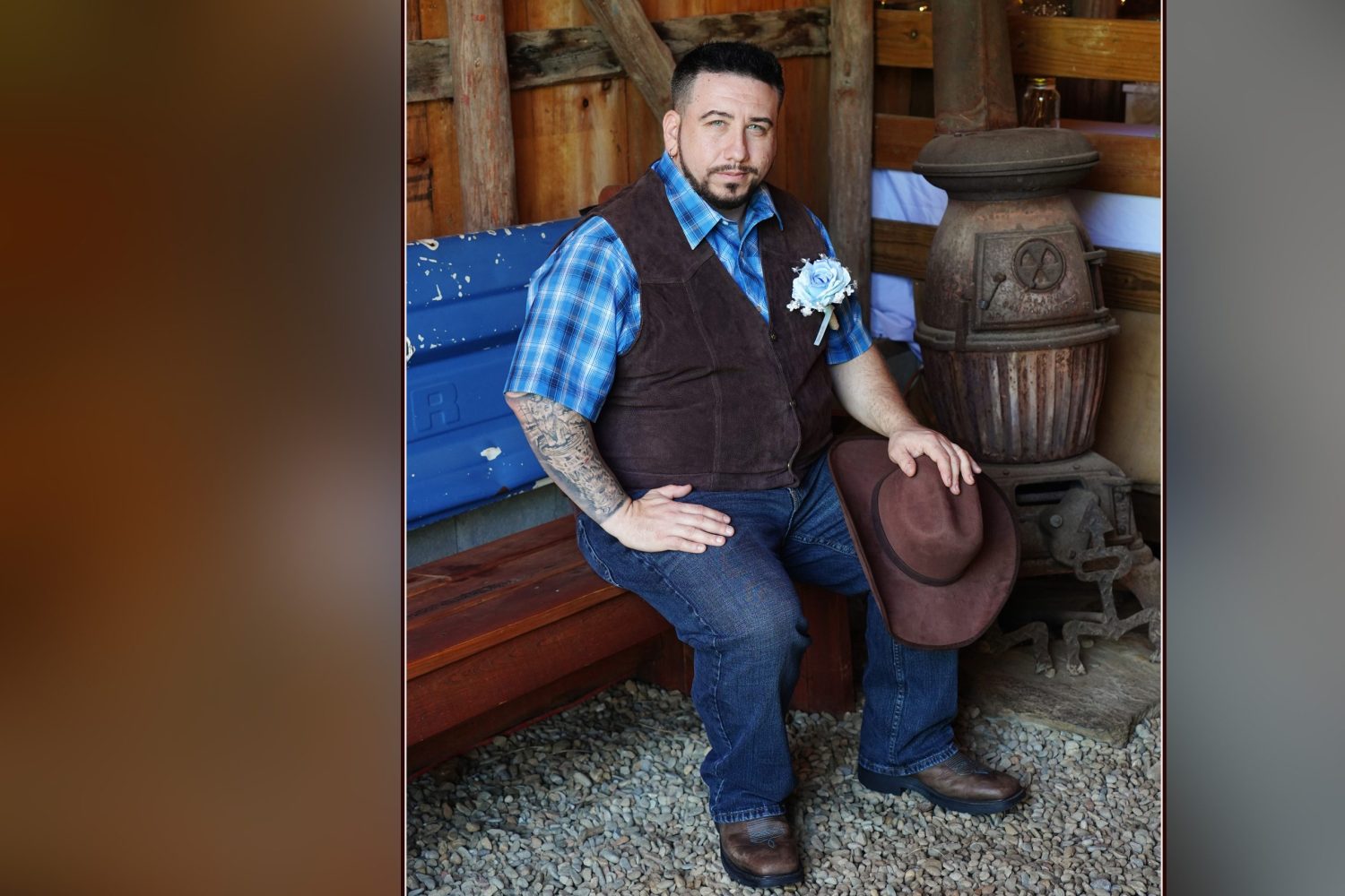 Groom with blue plaid and cowboy vest sitting by an antique pot belly stove holding his hat