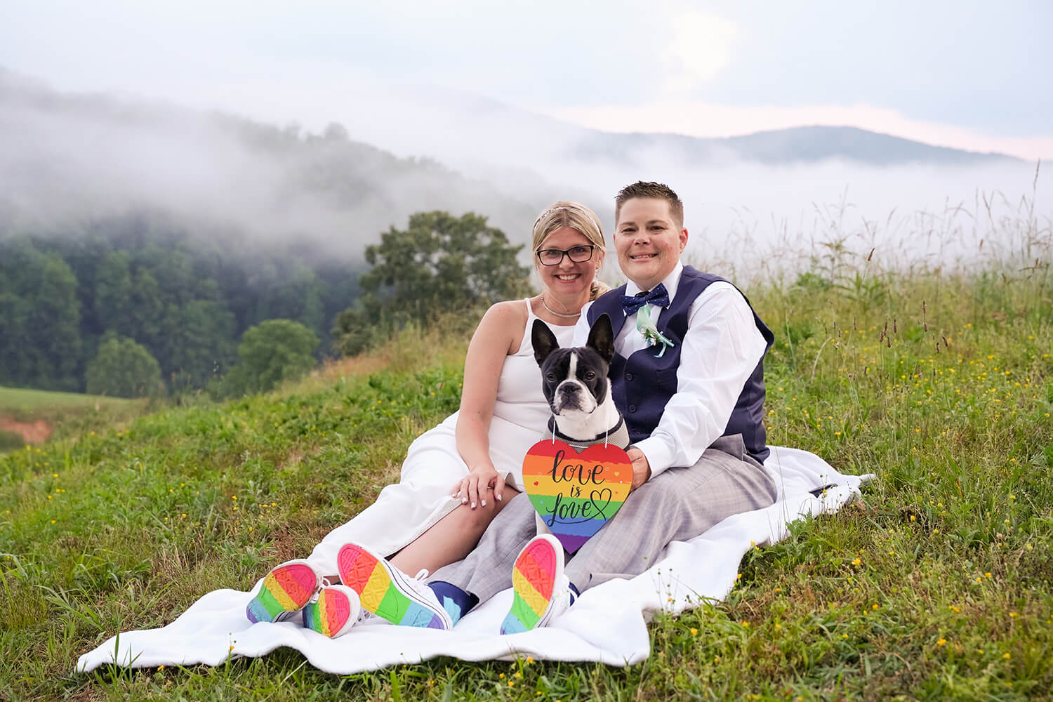 Two brides sitting on a blanket at the mountain view at Honeysuckle Hills after their same sex wedding with their Boston Terrior dog wearing a rainbow sign that matches the soles of their shoes and says Love is Love