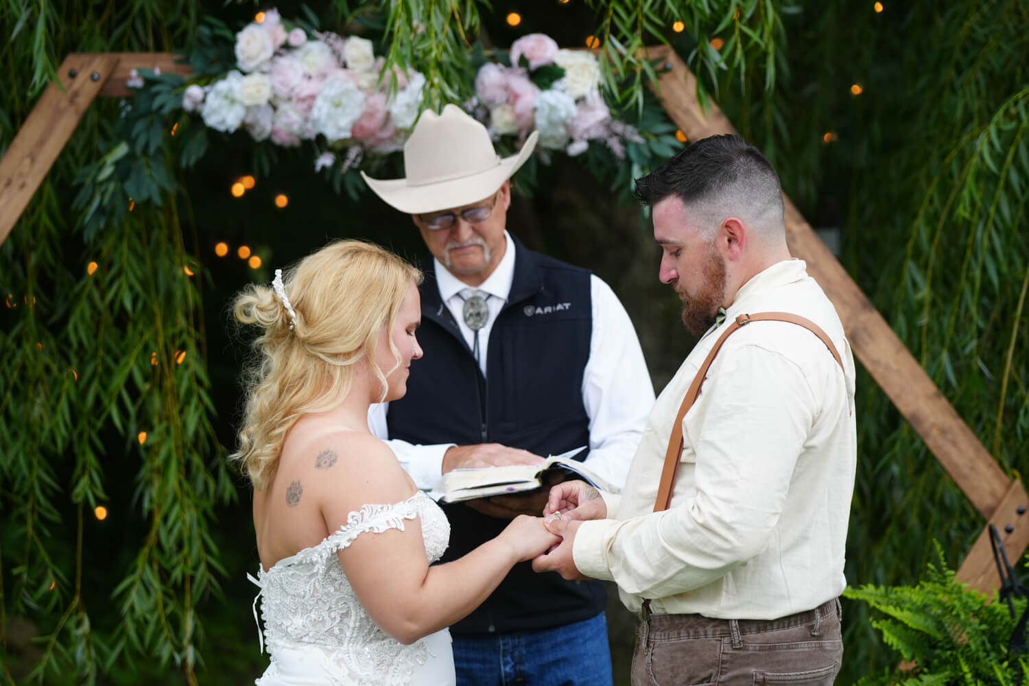 Pastor Lloyd officiating a wedding with his cowboy hat on at a hexagon arbor by a willow tree with lights as the couple exchanges wedding rings