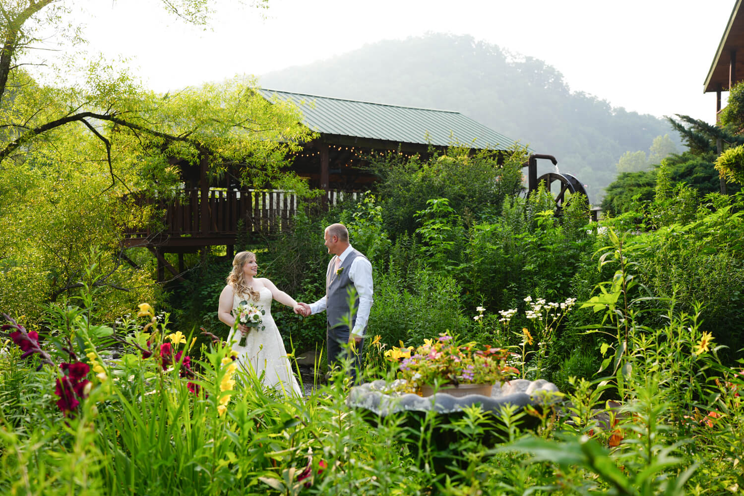 Couple holding hands on their wedding day in the summer flower garden at Honeysuckle Hills with a hazy mountain ridge and water wheel in the background