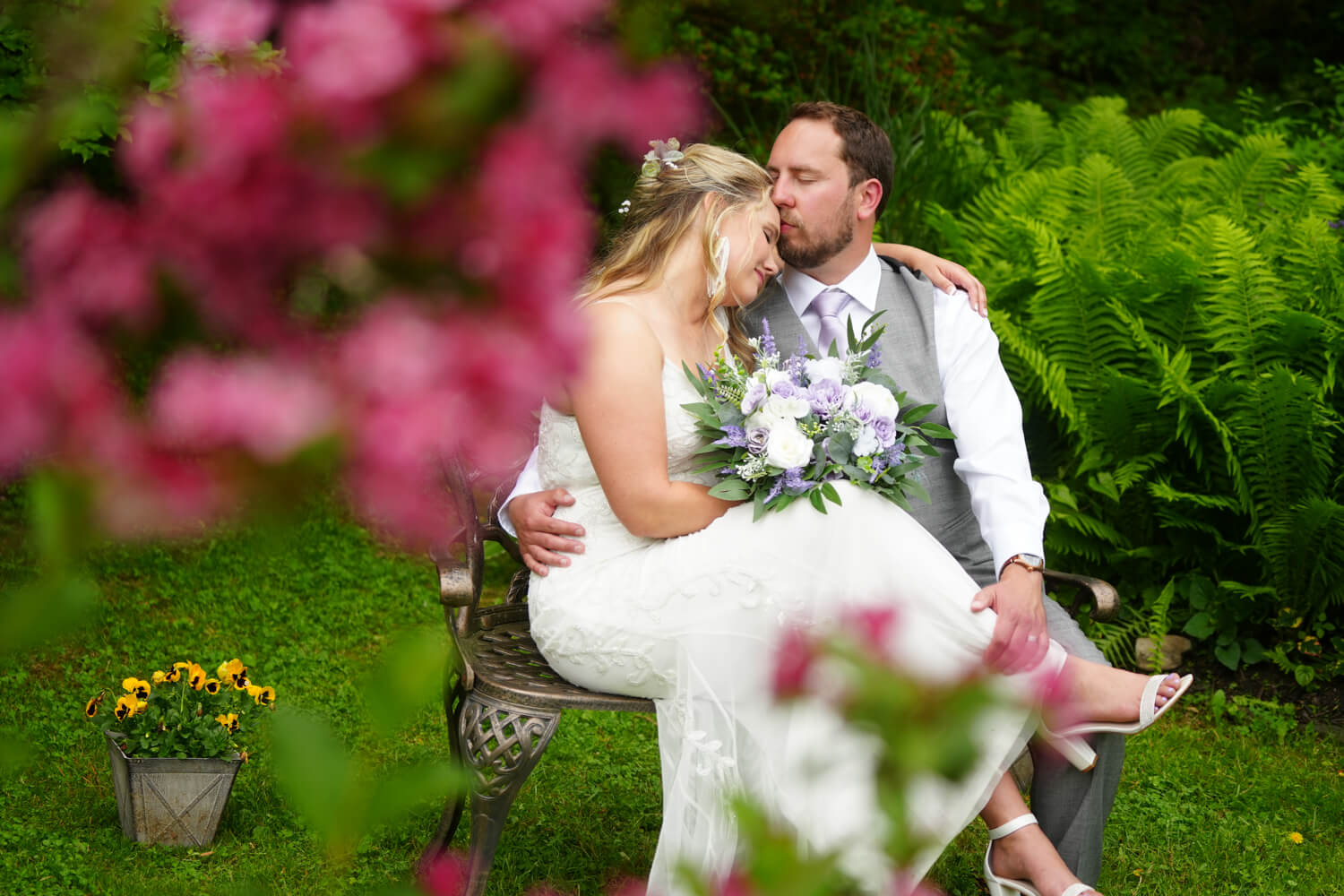 Bride sitting on her groom's lap in a spring flower garden with bright pink flowers and yellow pansies with ferns at Honeysuckle Hills near Gatlinburg TN