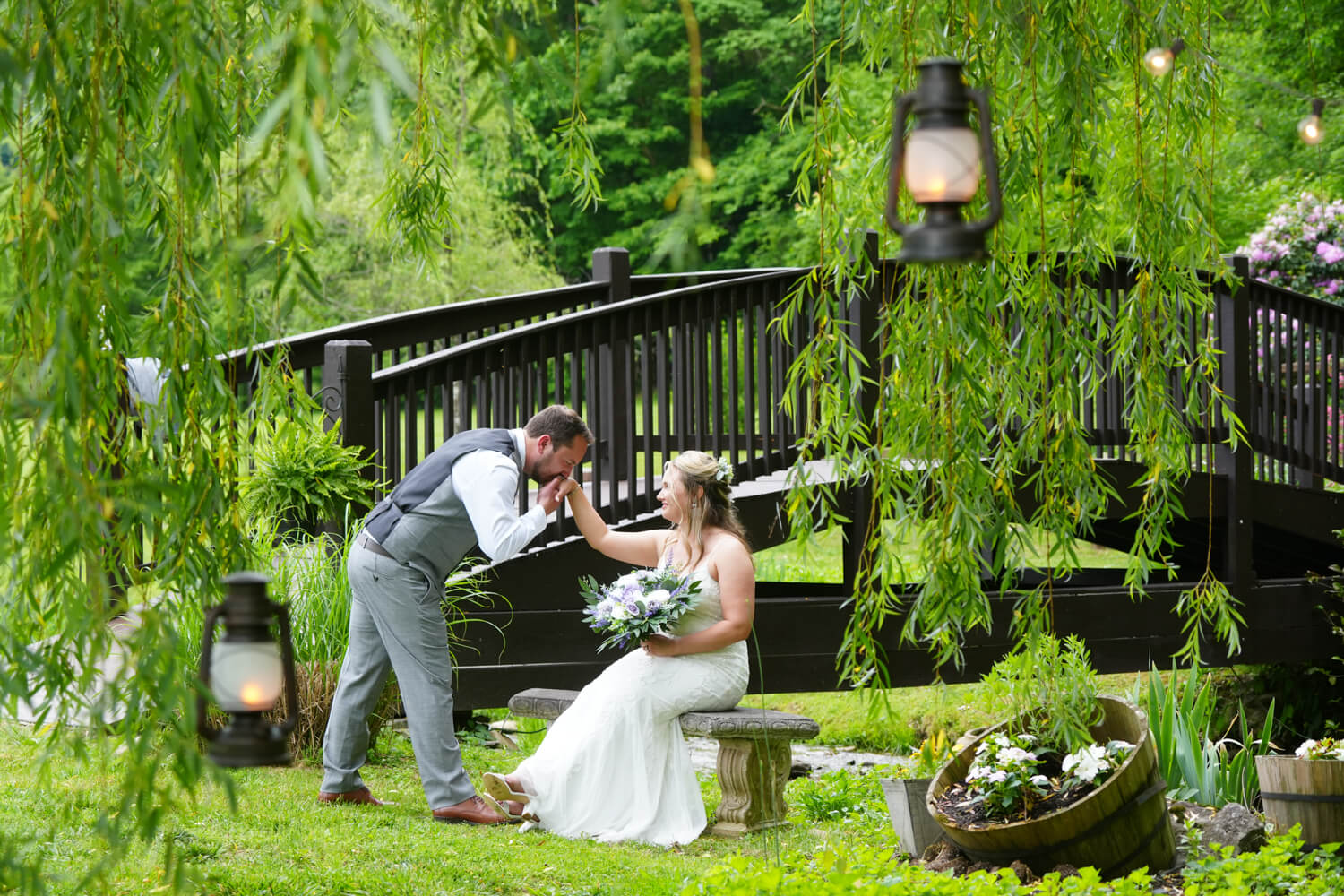 Groom kissing his bride's hand under a willow tree with hanging lanterns by the arched bridge at Honeysuckle Hills in Pigeon Forge