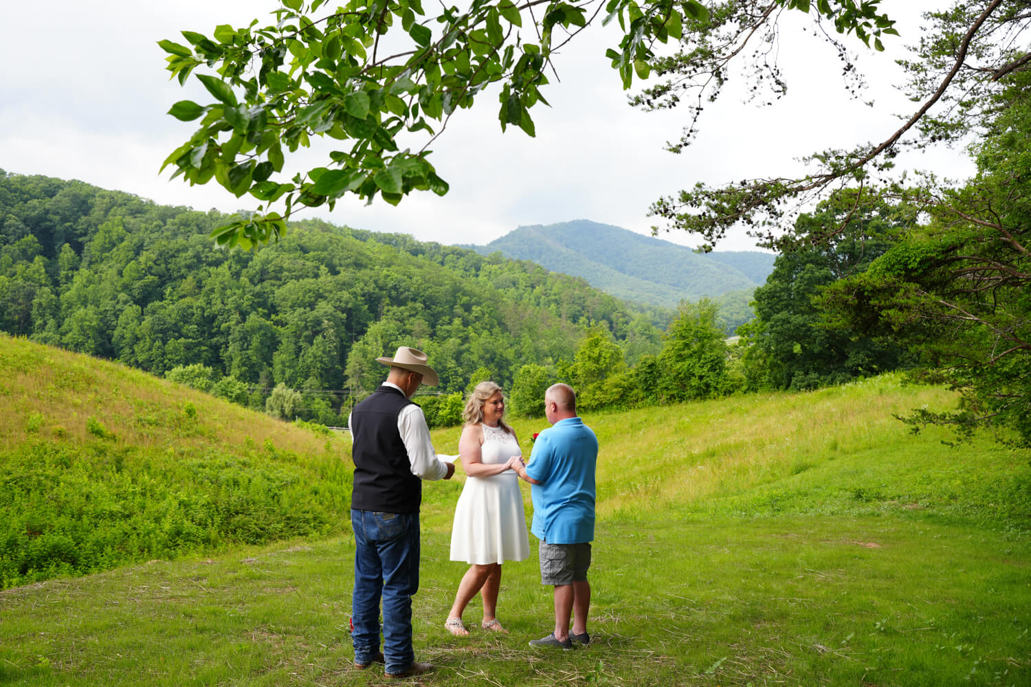 Pastor Lloyd officiating a ceremony in his cowboy hat for a casually dressed wedding couple at the mountain meadow view ceremony site at Honeysuckle Hills in Pigeon Forge