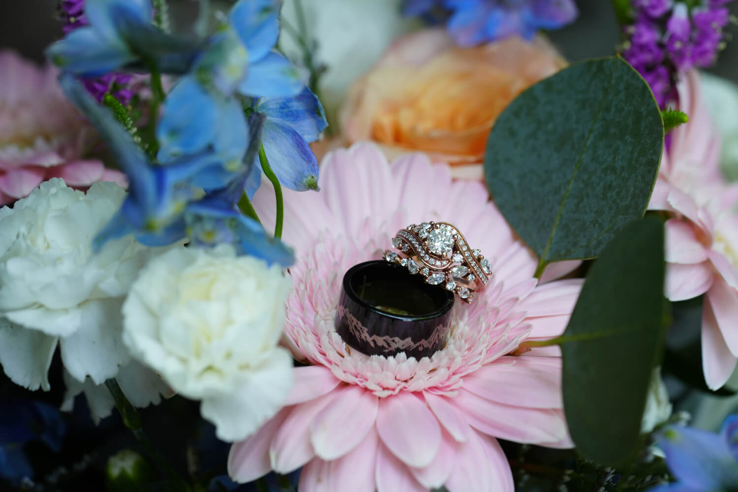 Ornate wedding rings sitting on pink and white flowers and the groom's ring has mountains on it