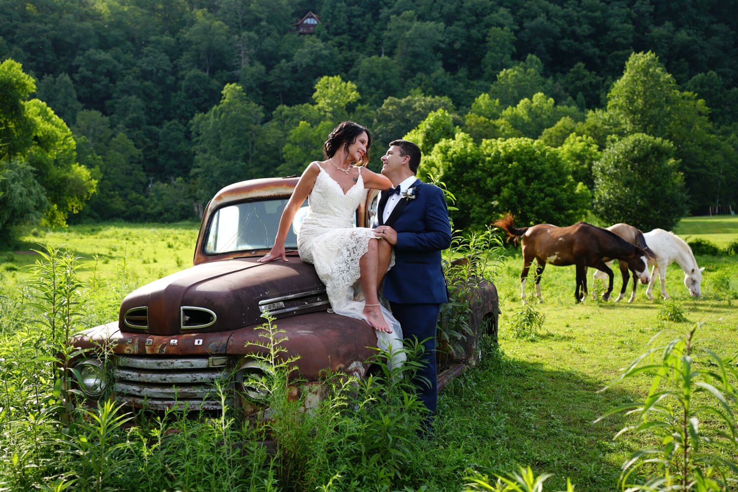 Bride in short white dress sitting on the hood of a 1950 Ford truck and leaning in to her groom with horses in the background