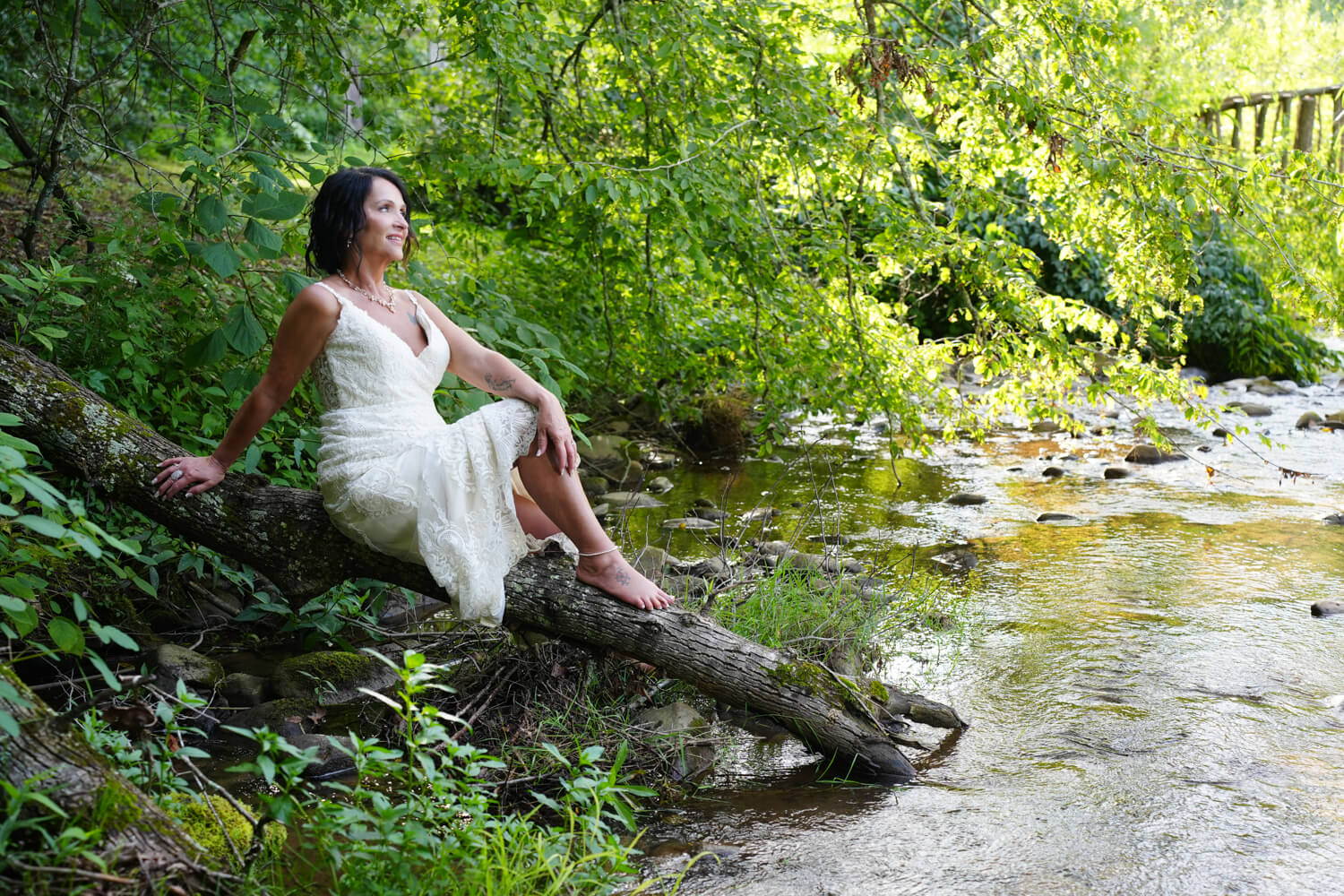 Bride in short white summer dress sitting on a log by a creek at the Honeysuckle Hills wedding venue in Pigeon Forge