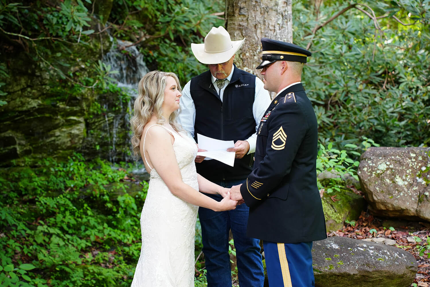 Groom in dress blues holding hands with his bride during the ceremony in front of a Smoky Mountain waterfall with a minister wearing a cowboy hat