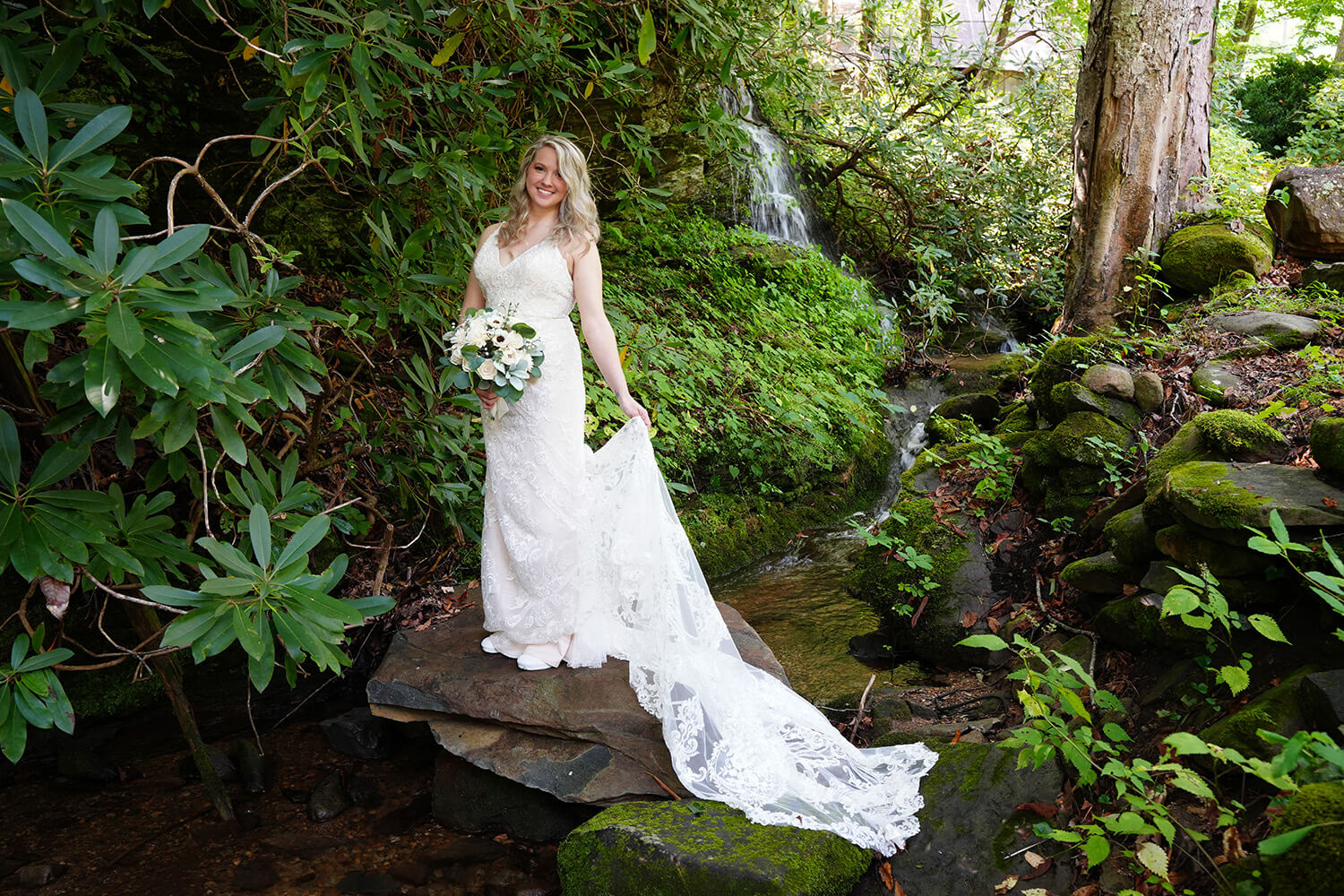 bride standing on a rock in front of a Smoky Mountain waterfall holding her long white train and a bouquet of white flowers