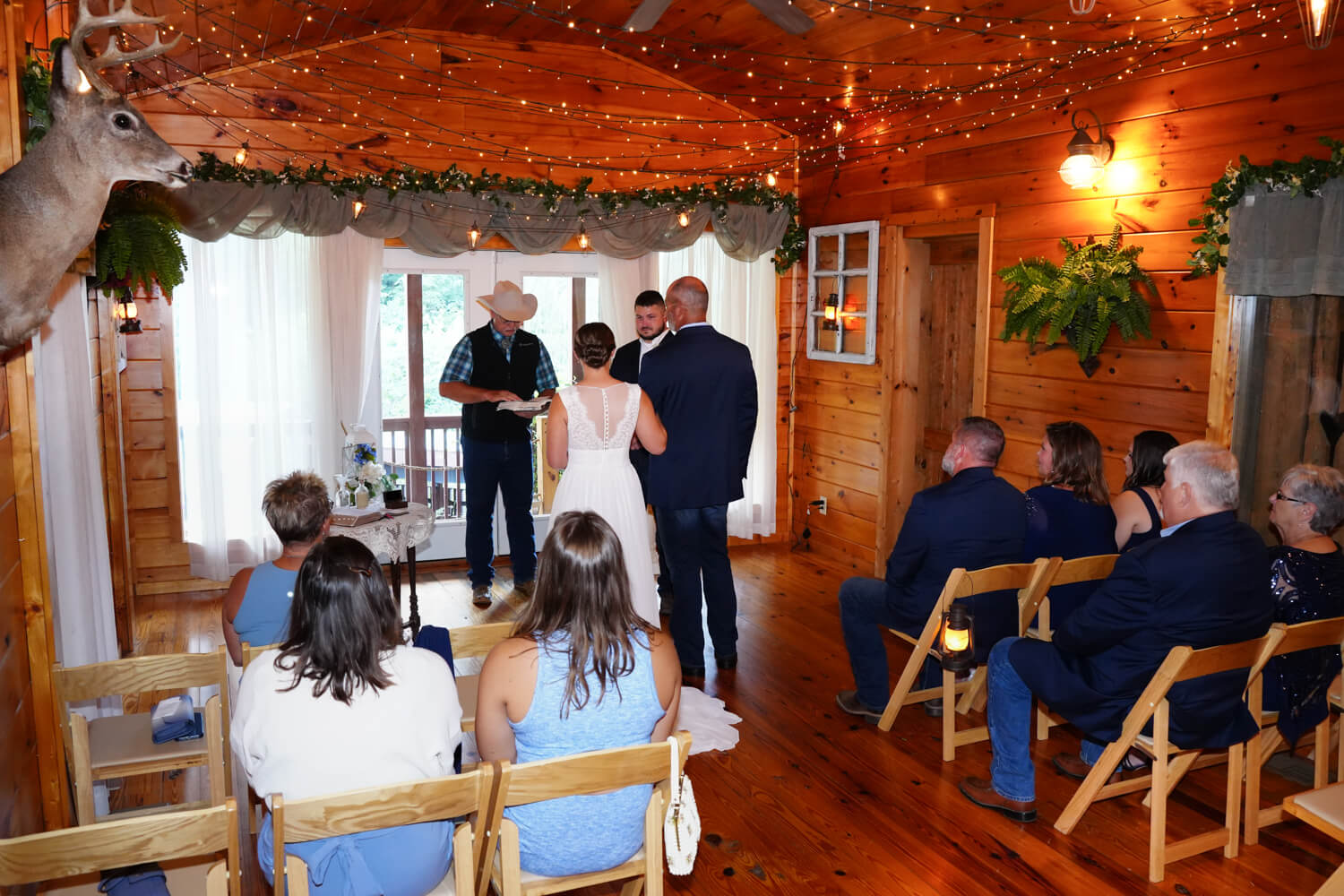 Tennessee Wedding Chapel with wooden pine walls, fairy lights, lanterns and a deer head on the wall as guests seated in wooden chairs watch a preacher with a cowboy hat ask the father who gives the bride away