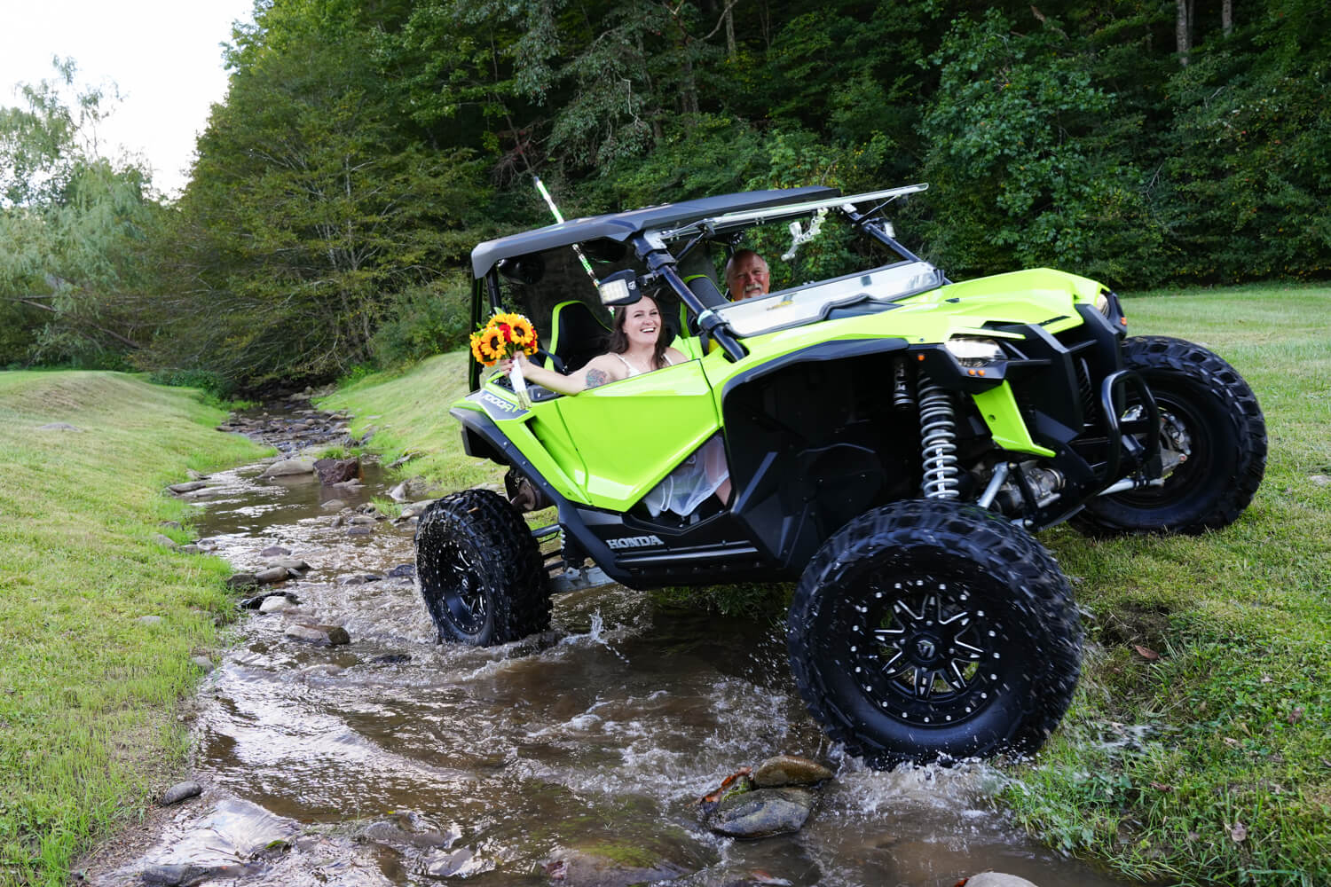 Bride waving her bouquet outside the window of a neon green side by side all terrain vehicle driving through a creek in the Smoky Mountains