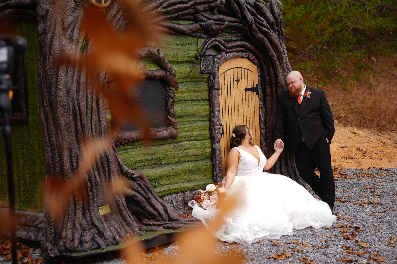 Yellow fall leaves falling in front of a green fairy house with as a groom holds the hand of his bride seated at the door