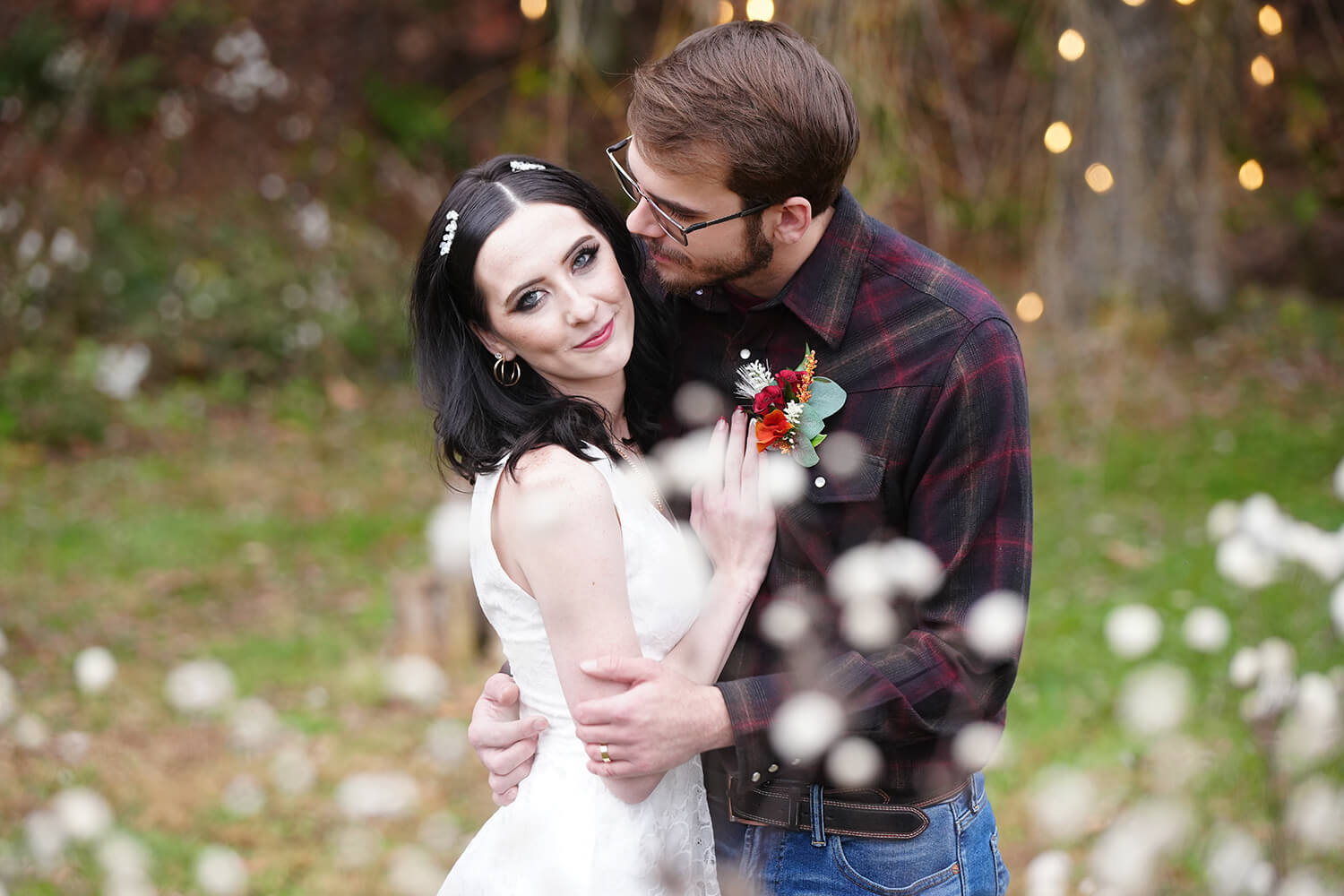 Bride standing in front of white cottony flowers with jet black hair and piercing blue eyes leaning in to her groom in a plaid shirt who wants to kiss her cheek