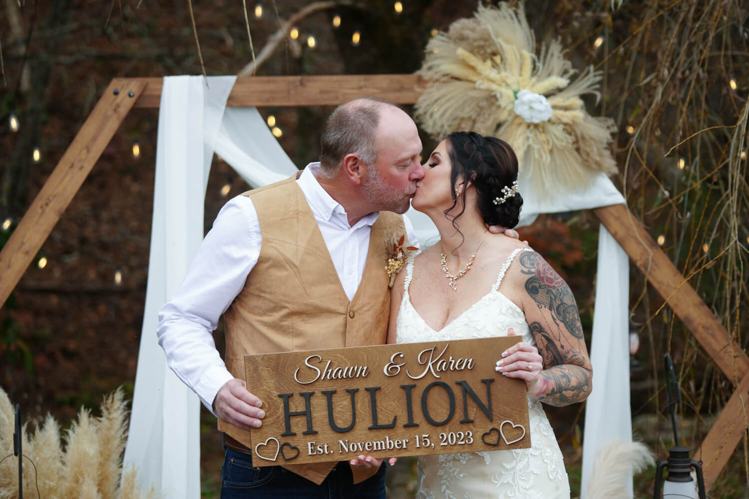 Couple kissing in front of a hexagon boho arbor while holding a sign with their last name Hulion