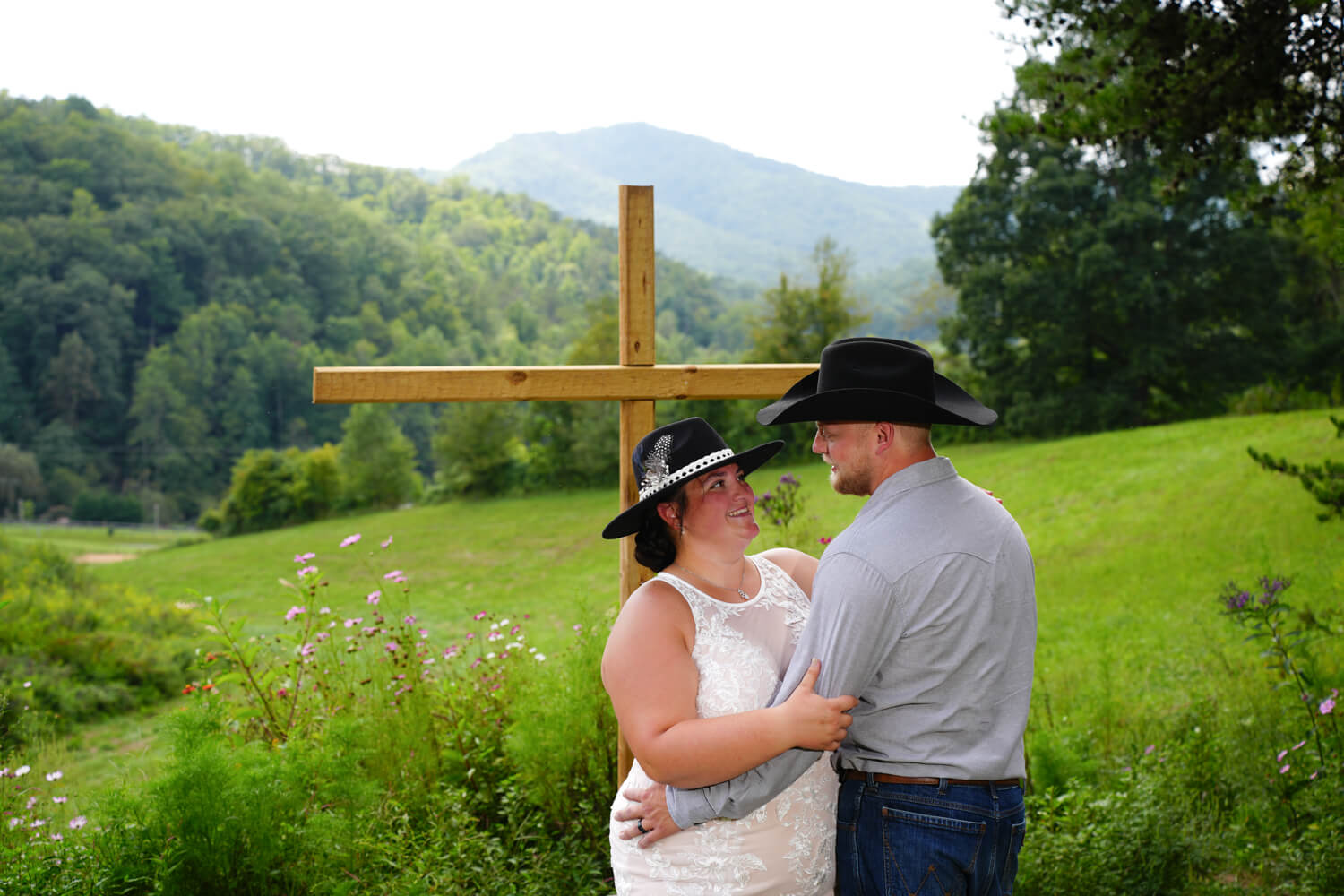 Western wedding couple with black cowboy hats posing for a picture at a wooden cross in a meadow with a mountain ridge view in Pigeon Forge, Tennessee