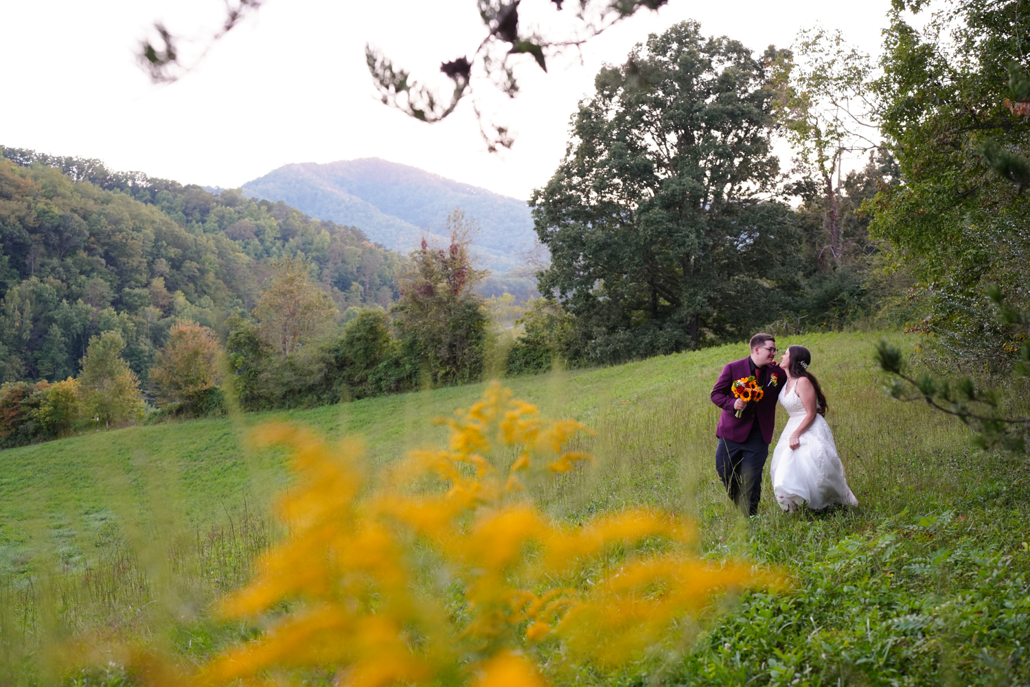 Groom holding his bride's bouquet as the walk and laugh in a meadow with golden rod and a mountain view