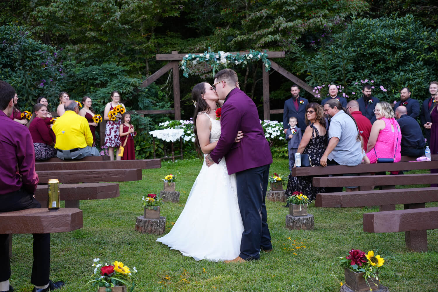 Bride and groom kissing in front of a western style wooden arbor at the base of a mountain ridge as their guests watch