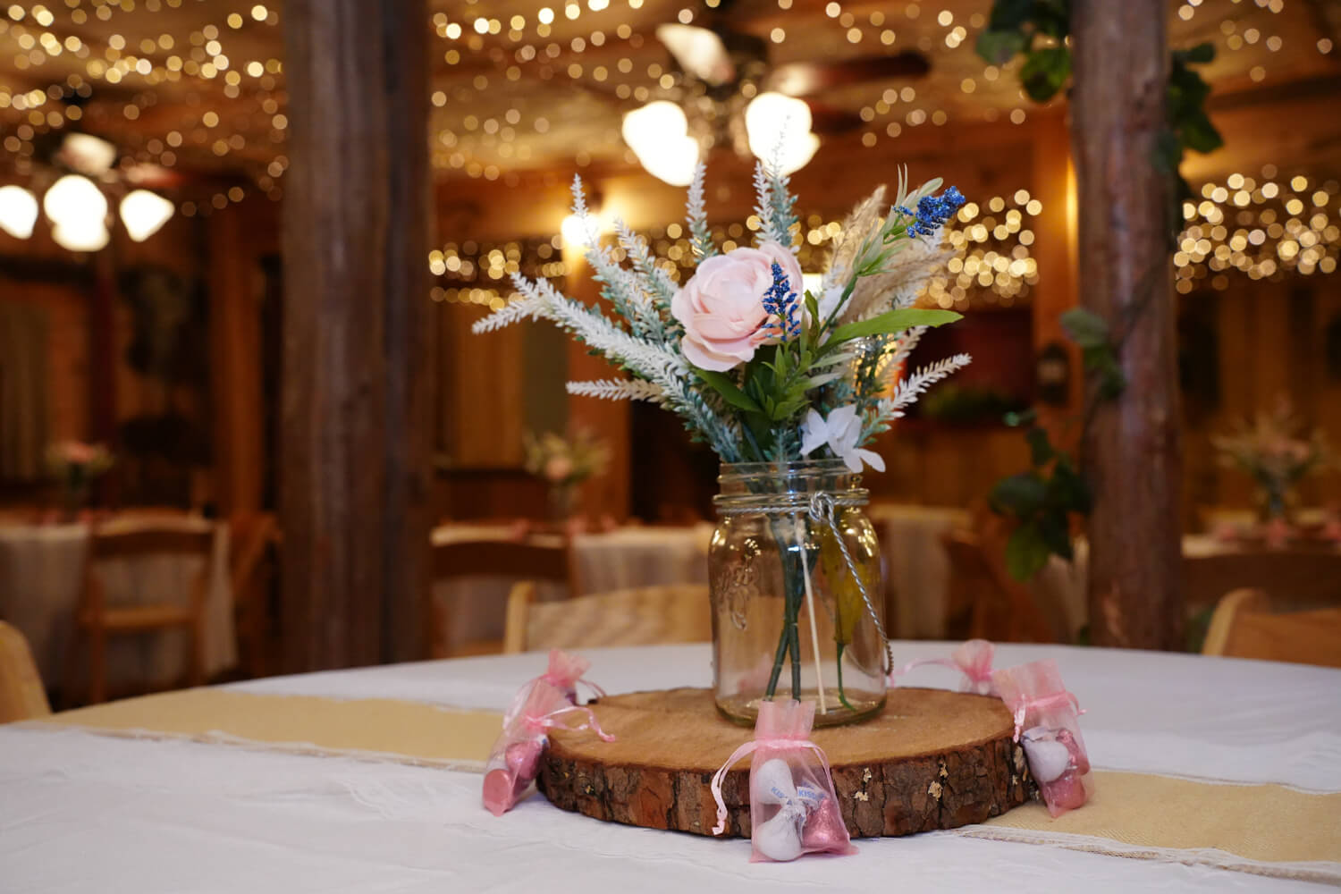Glass vases with greenery and pink flowers sitting on a log slice on a reception table in a country chic barn wedding venue