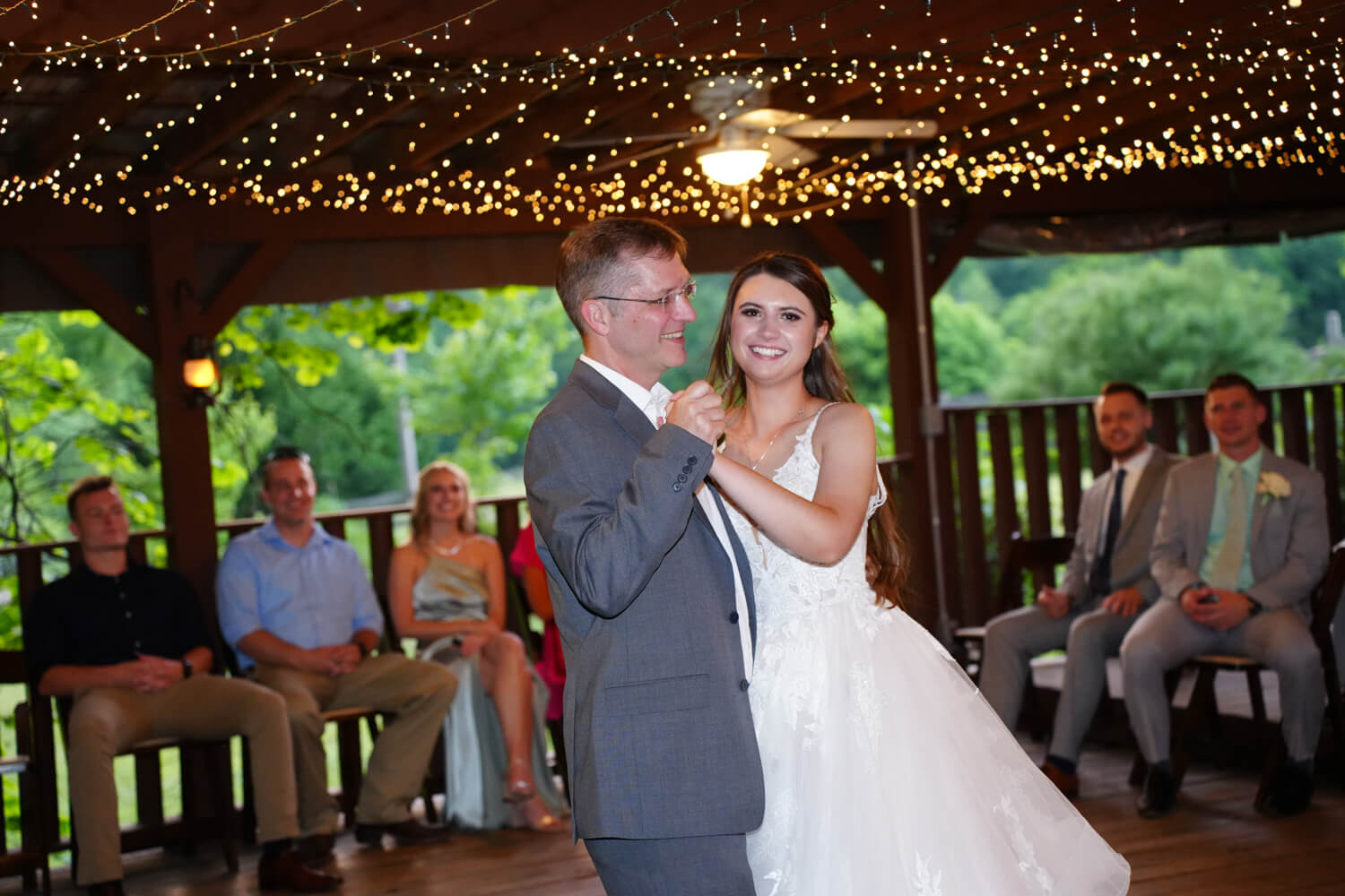 Father daughter dance on wedding day in a pavilion with pretty fairy lights on the ceiling and guests watching in the background