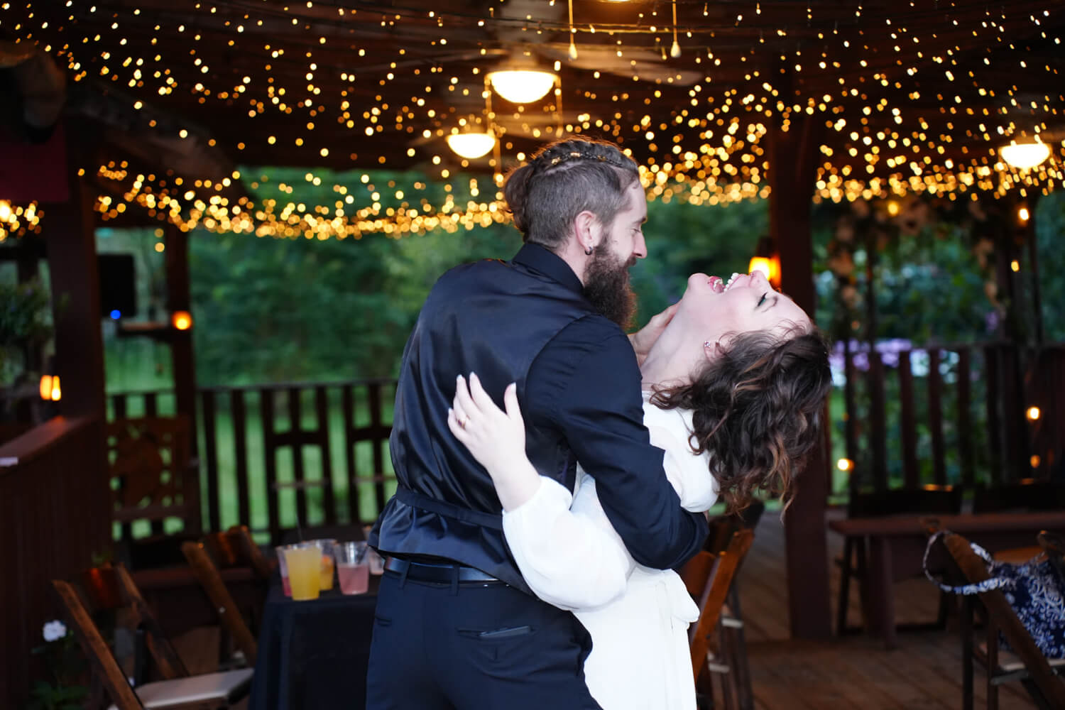 Groom dipping his bride beneath warm fairy lights on the ceiling of a creekside pavilion at their wedding reception