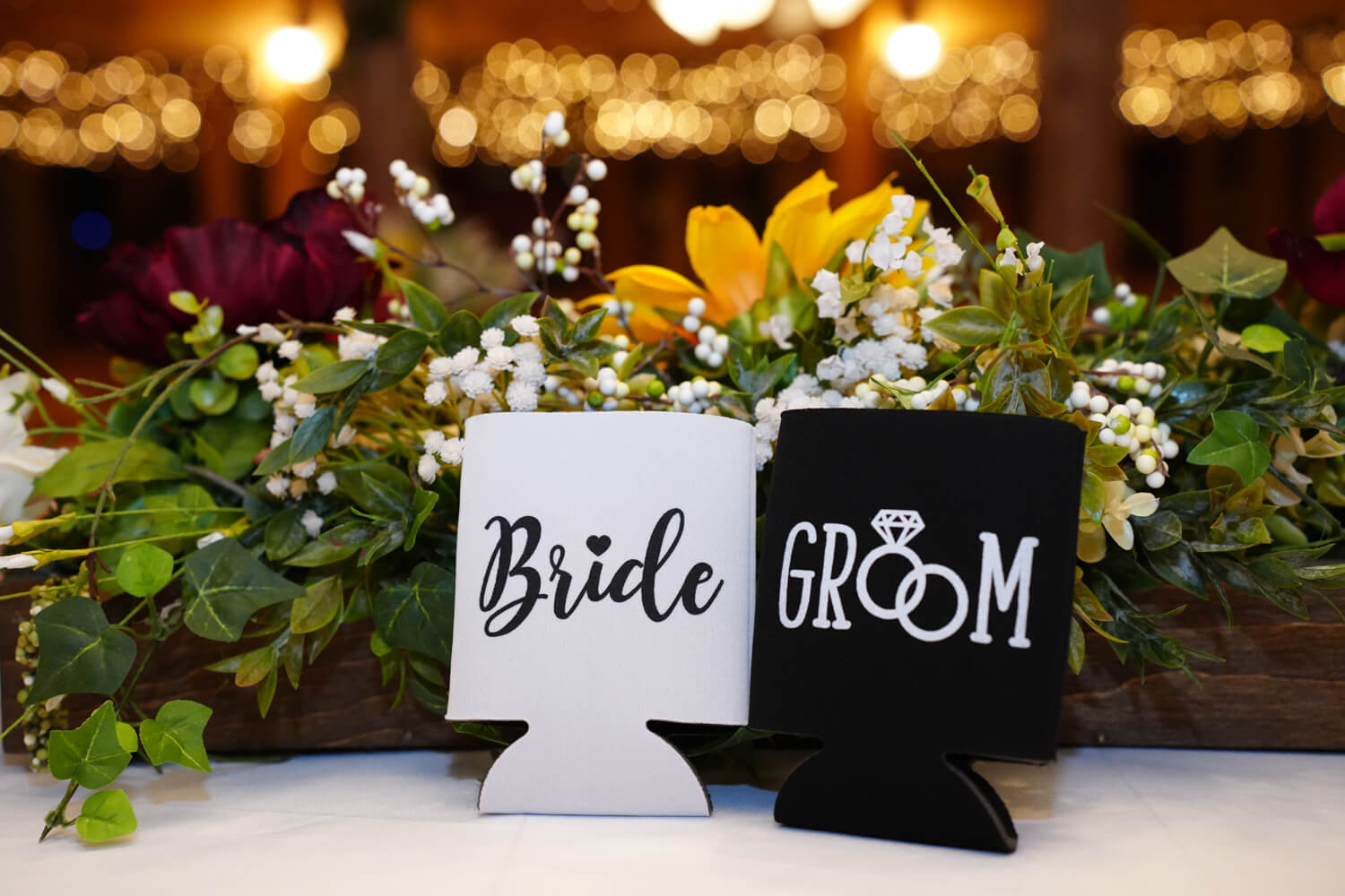 Bride and groom koozies sitting on a table with white flowers and warm string lights in the background