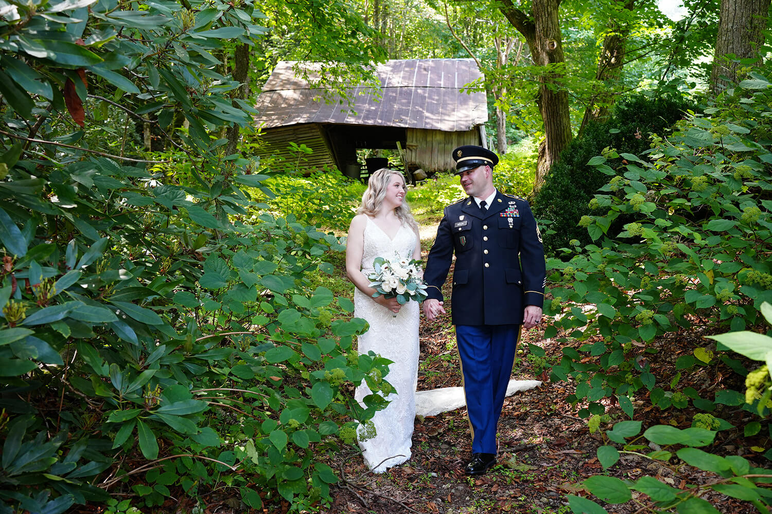 Bride walking holding hands with her groom in military dress blues on their wedding day down a forest path with a historic barn behind them