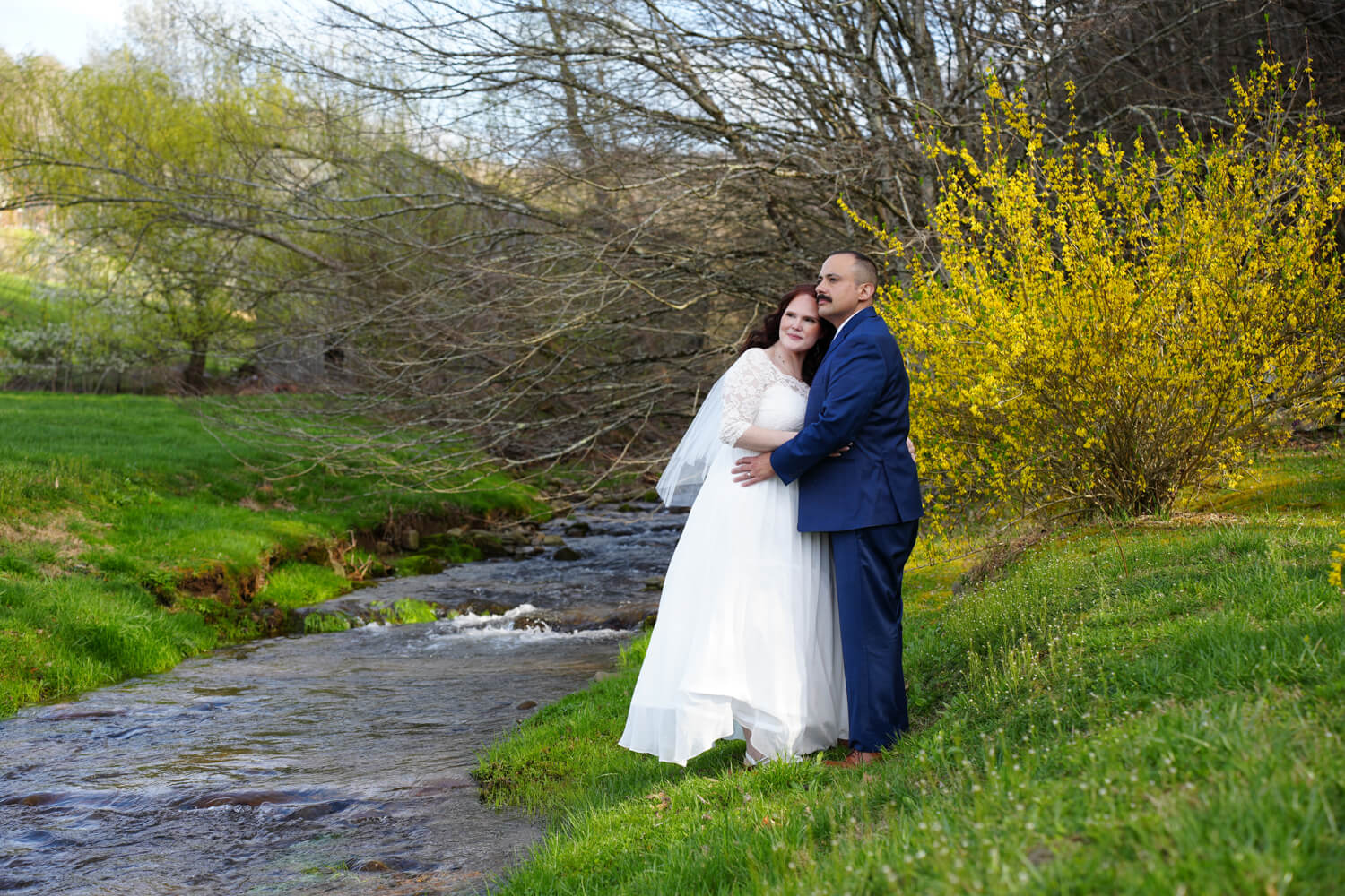 Bride in a white wedding dress and white veil leaning closely to her husband's cheek by a creek in early spring by a large blooming yellow forsythia bush at the Honeysuckle Hills Wedding Venue in Pigeon Forge Tennessee