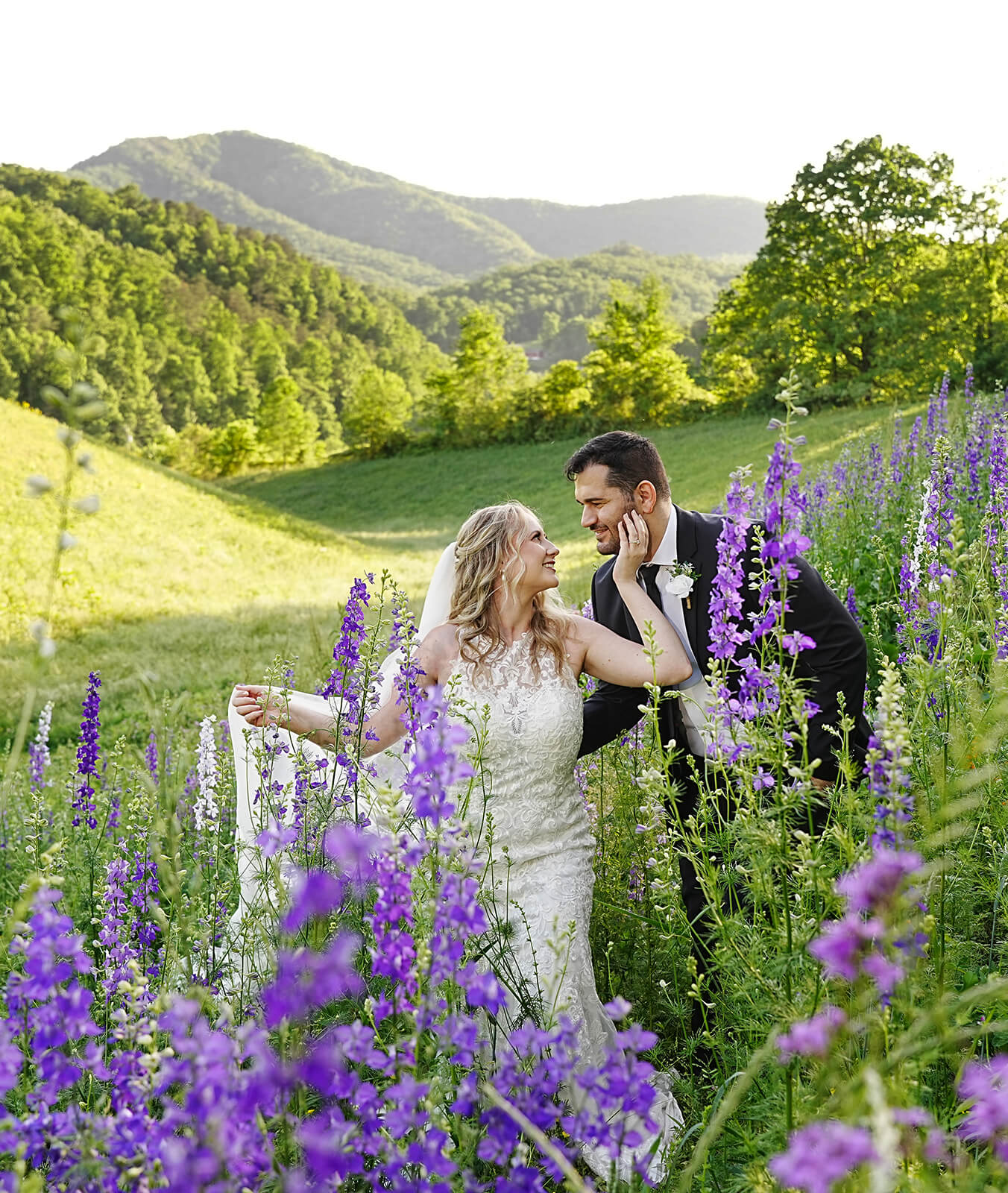 Bride holding her veil and touching her husband's cheek in a field of purple larkspur wildflowers with a mountain view behind them