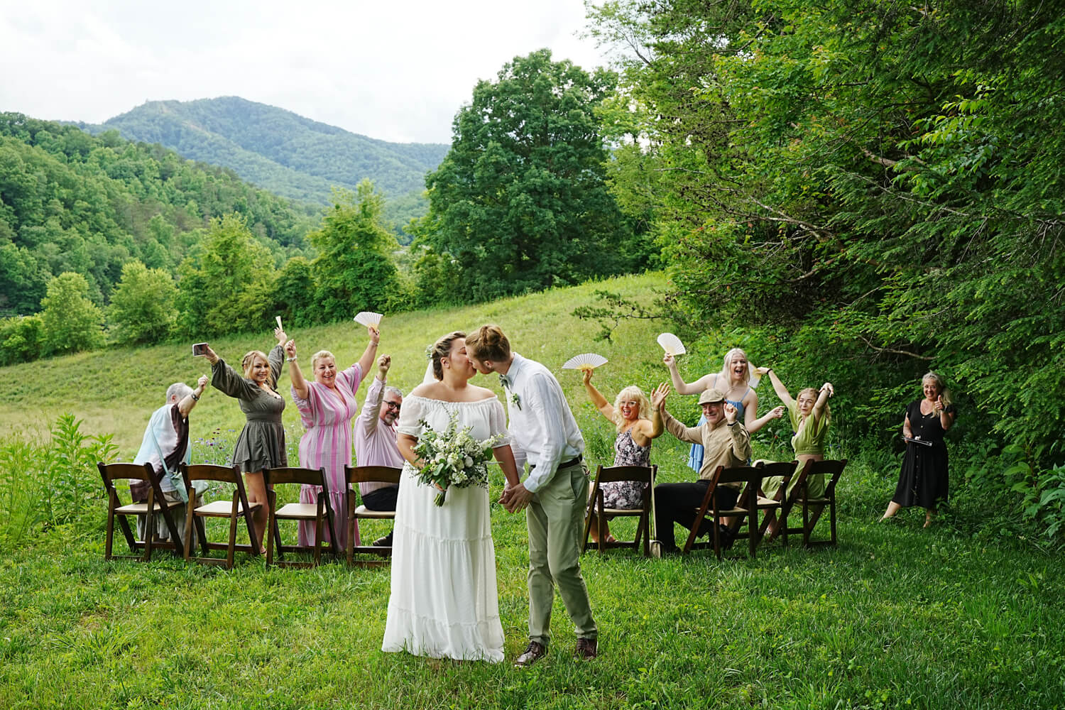 Wedding guests waving their hands in celebration as the couple kisses in front of a mountain view ceremony site