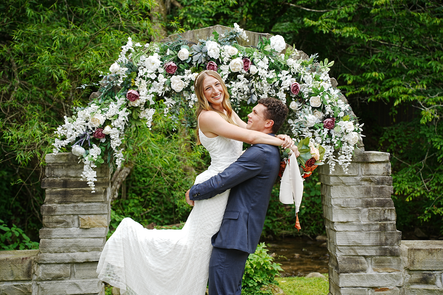 Groom lifting up his bride in a white sun dress on their wedding day at a stone arch covered in white and pink flowers