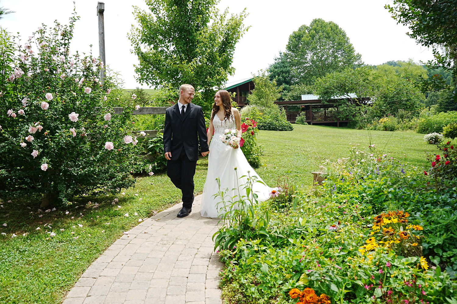Couple walking down a brick path in a summer garden with a barn behind them on their wedding day