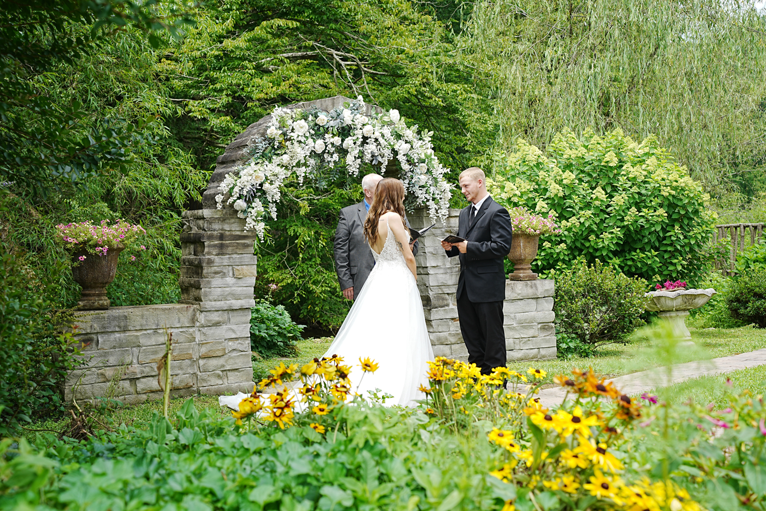 Micro wedding ceremony at a stone arch decorated with white flowers in a summer garden with blooming yellow flowers