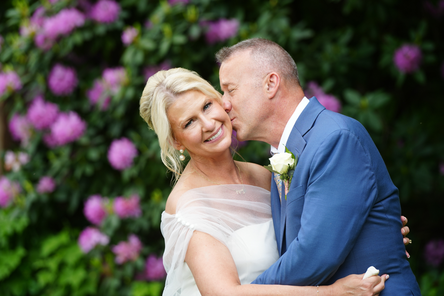 middle aged groom kissing his bride on their wedding day with purple rhododendron in bloom