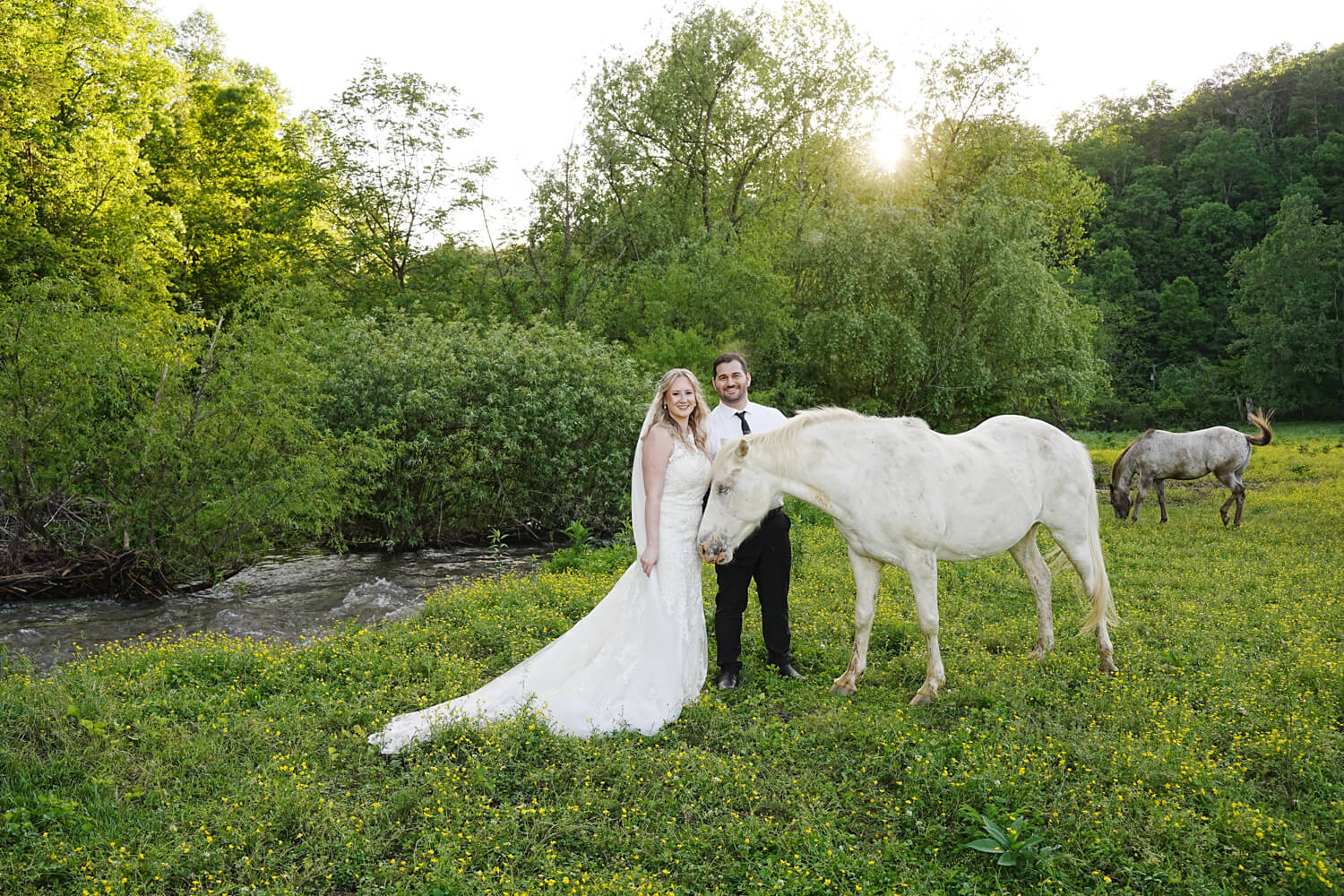bride and groom petting a white appaloosa horse in a field by a creek as the sun goes down behind the trees