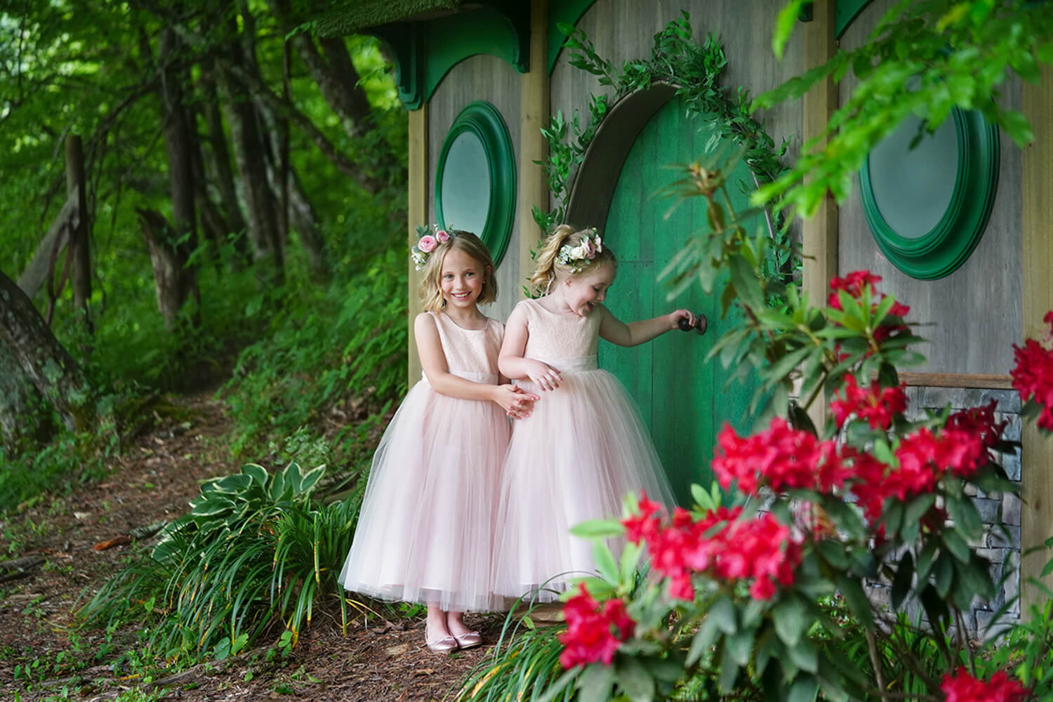Two flower girls in pink dressing at the round green door of a fairy hobbit house with bright pink flowers in bloom