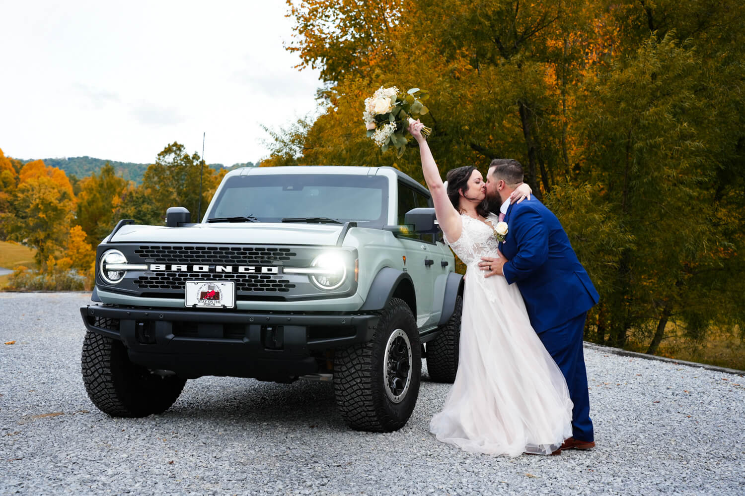Bride holding up her bouquet in joy as her groom kisses her by their new Ford Bronco in the fall in the Smoky Mountains